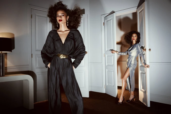 Two women in elegant evening wear in an interior setting. One wears a black jumpsuit, the other wears a silver dress emerging from a door.