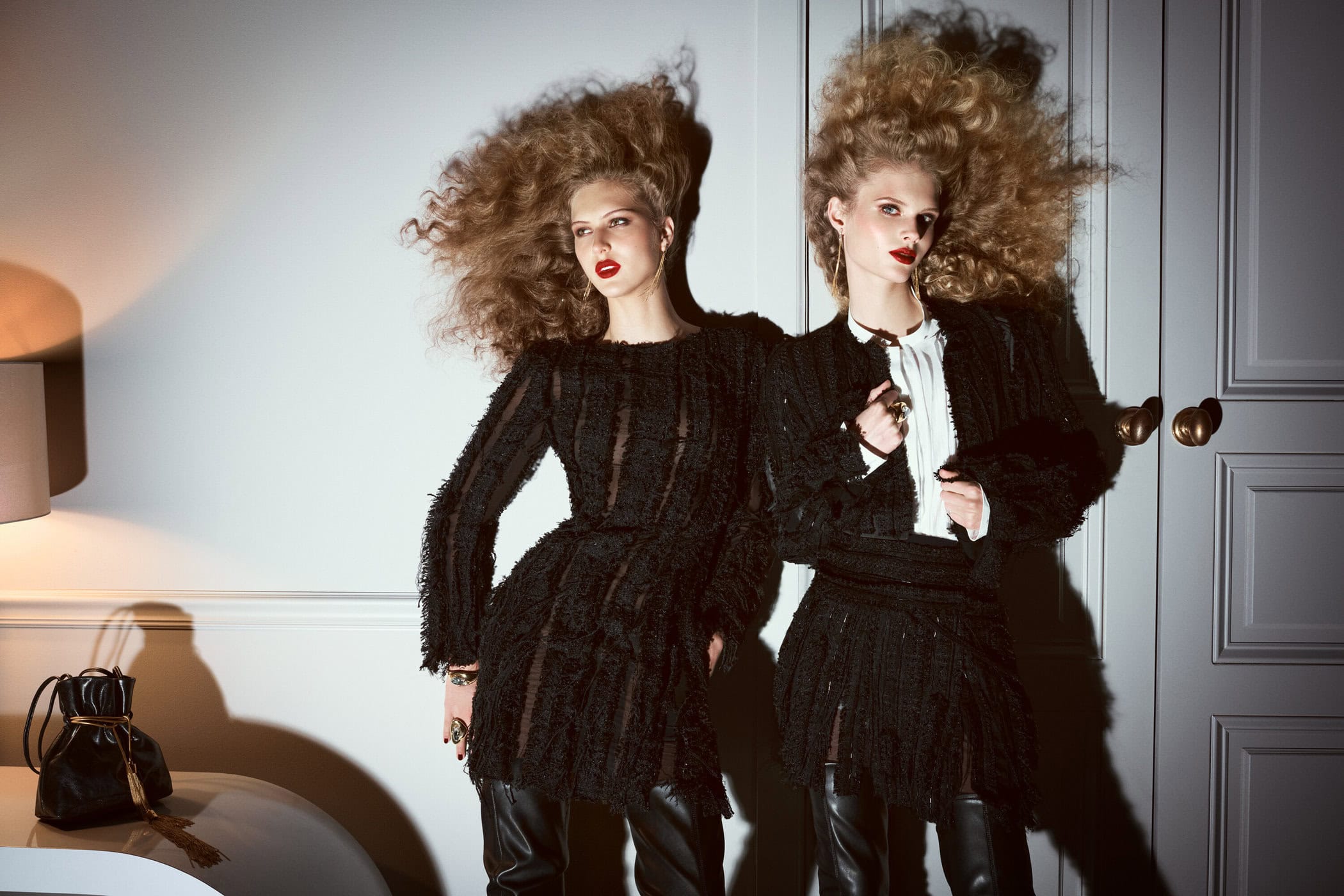 Two fashion models with curly hair pose indoors in black dresses and leather boots, accessorized with jewelry, in a studio setting.