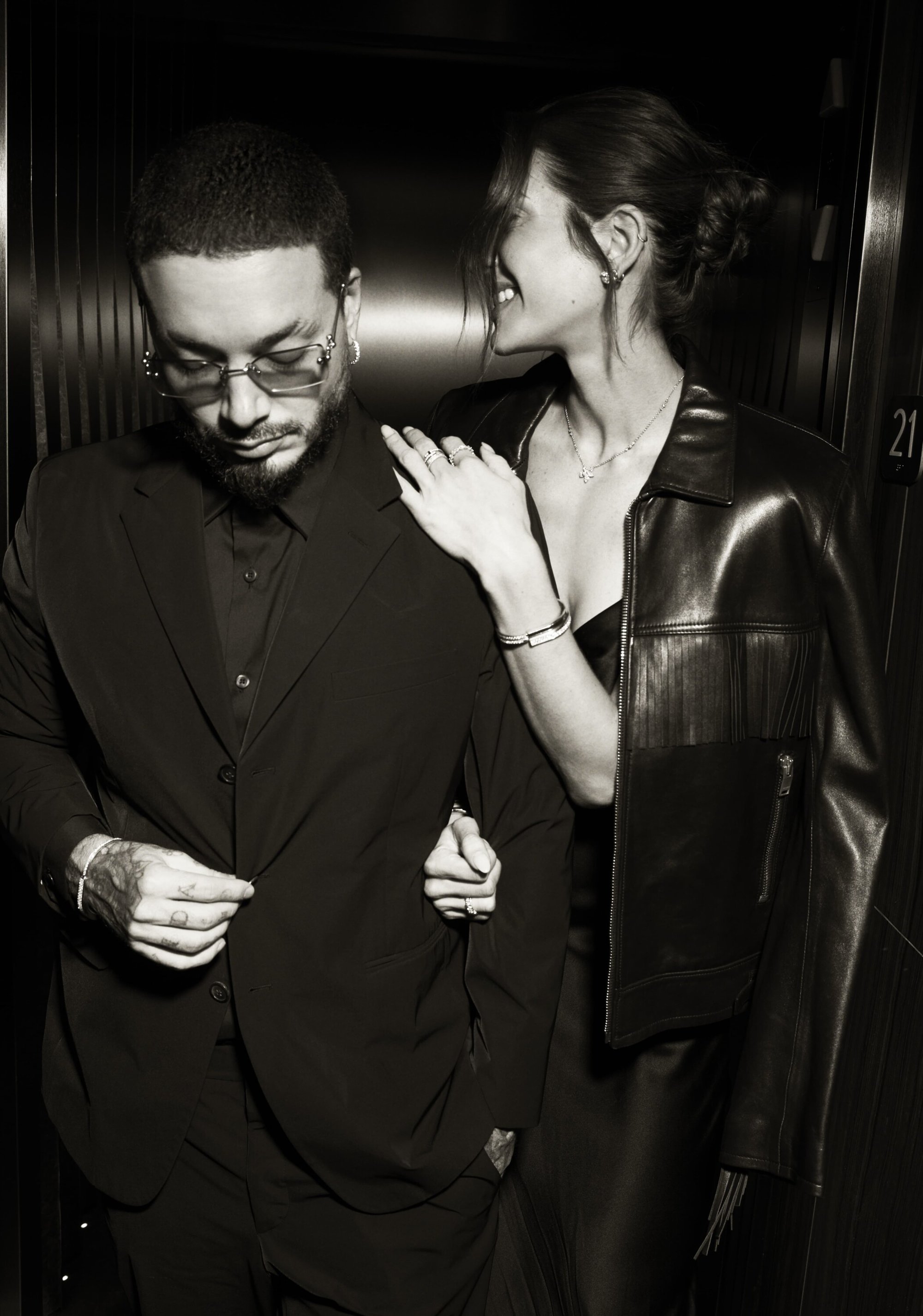 Monochrome photo of a stylish couple posing indoors; the man in a suit and glasses, the woman in a leather jacket looking at him.