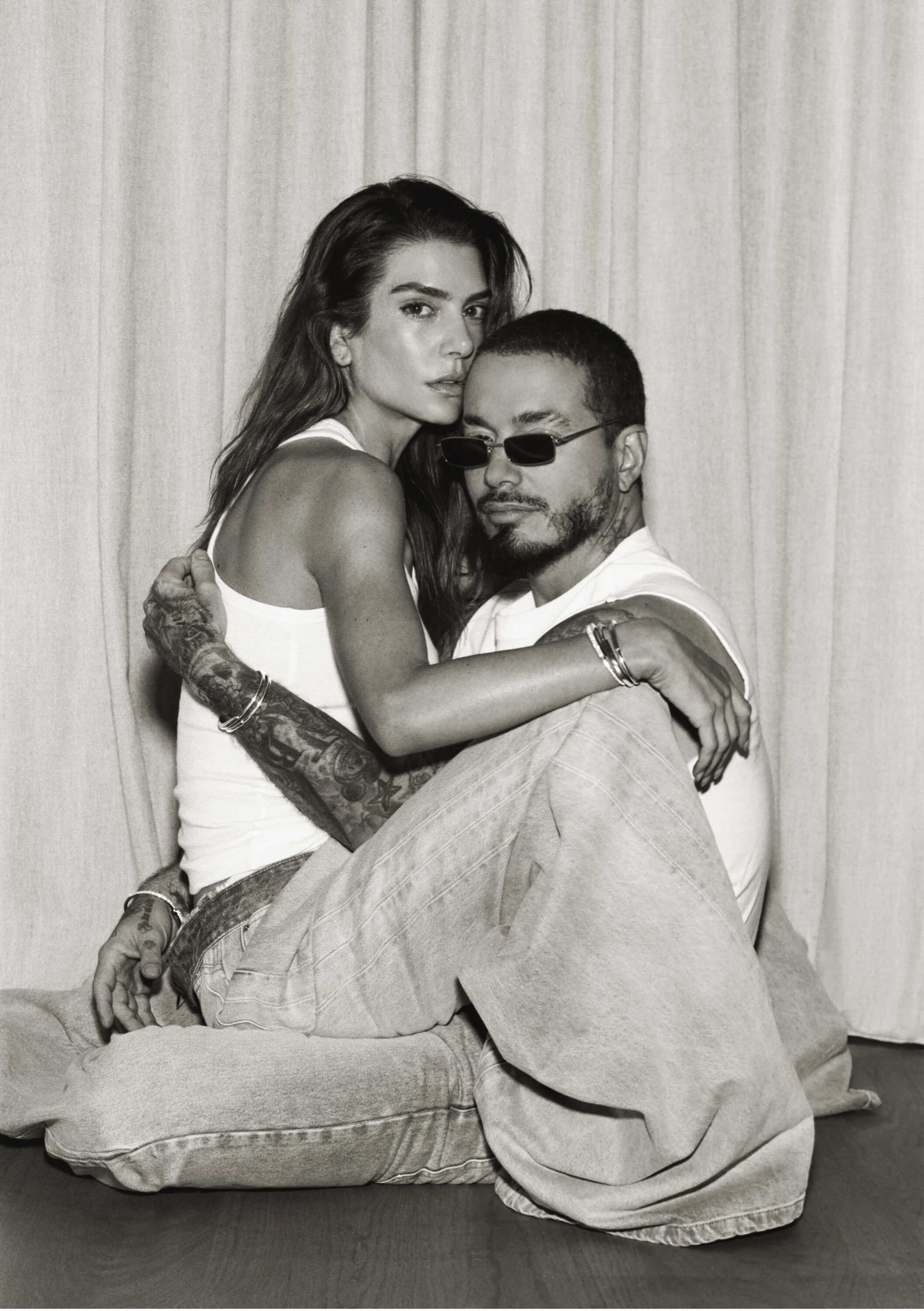 Monochrome portrait of a woman in a white tank top embracing a seated man with tattoos and sunglasses in front of a curtain backdrop.