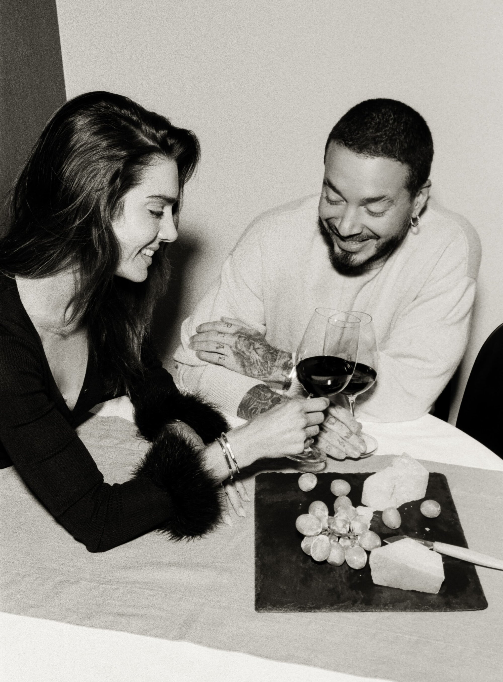 Woman and man share wine at table with grapes and cheese in a black-and-white photograph showcasing an indoor setting and joyful expression.