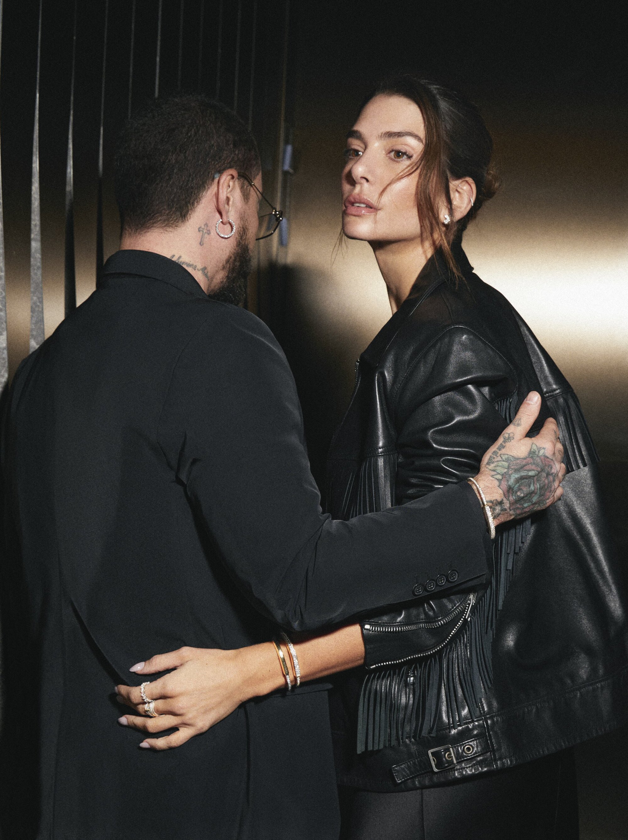 Stylish couple poses indoors: adult woman in a leather jacket with fringe detail alongside a man in a dark suit.