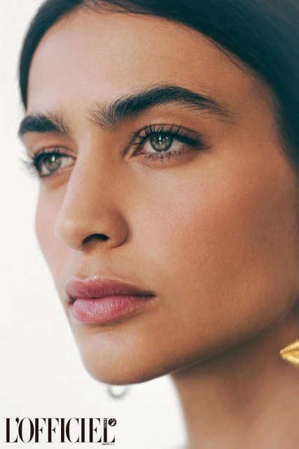 Close-up portrait of a young woman with green eyes and tan skin. She has thick eyebrows, pink lipstick, and gold earrings.