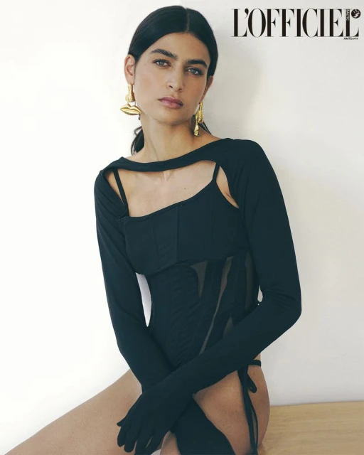 A woman poses in a black bodysuit and gold earrings in a studio, looking at the camera with a neutral expression in soft light.