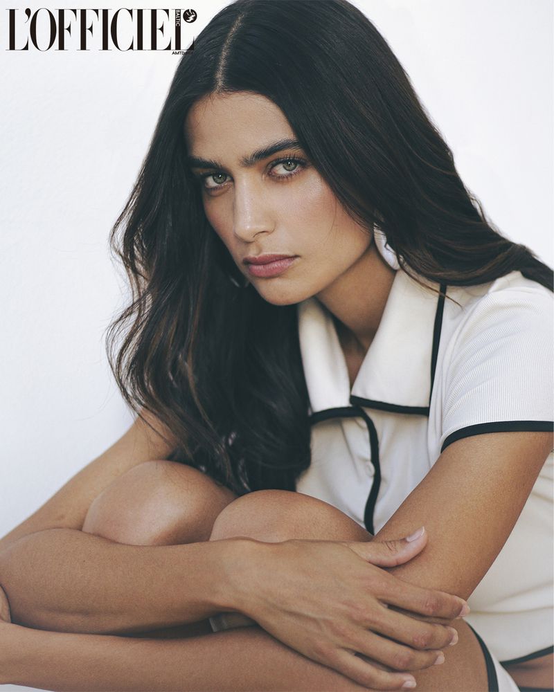 Close-up studio shot of a young adult woman with long dark hair wearing a white top with black trim, gazing intently at the camera.