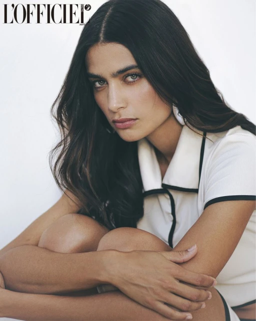 Close-up studio shot of a young adult woman with long dark hair wearing a white top with black trim, gazing intently at the camera.