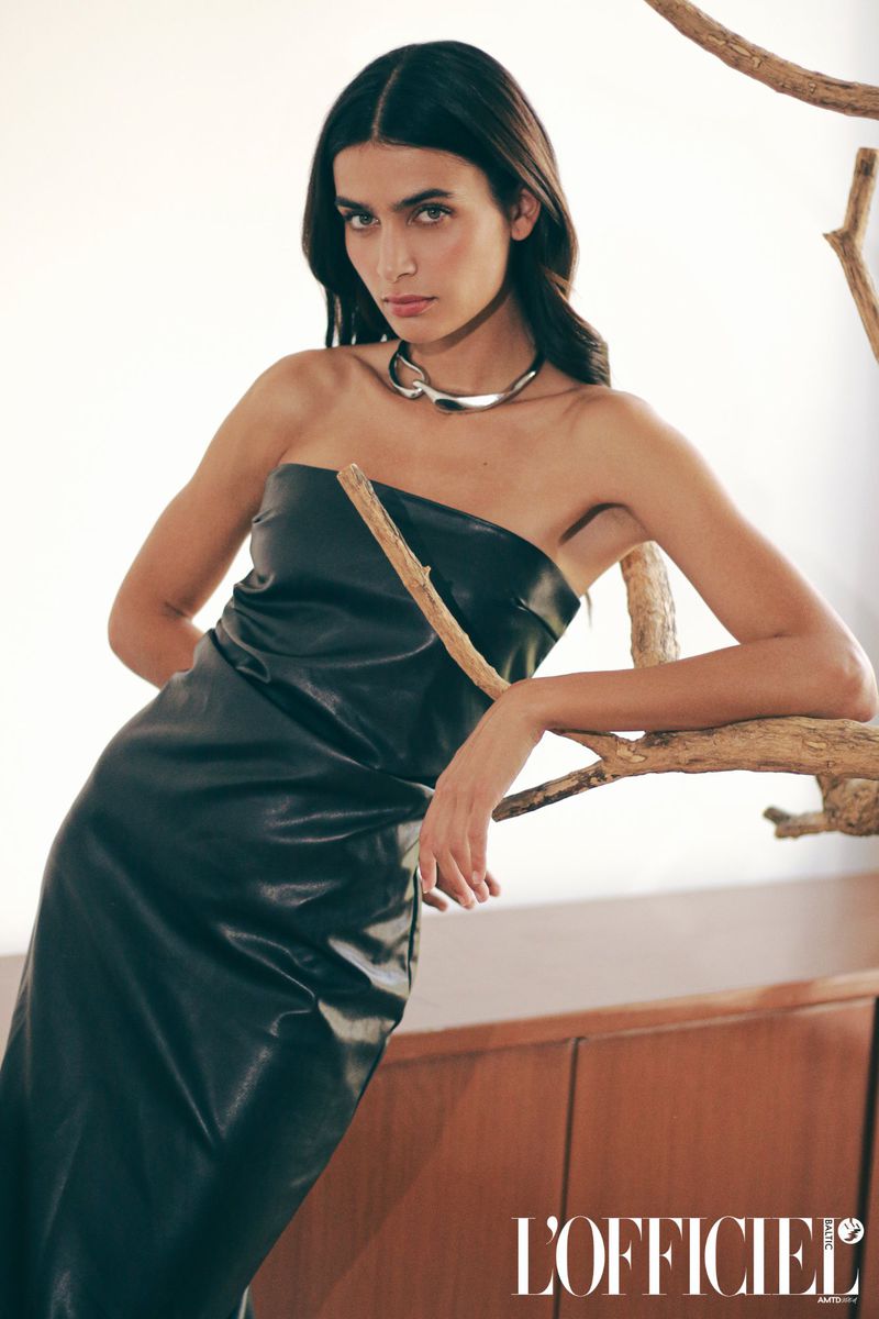 Brunette woman in a black, strapless, shiny dress with a metal necklace, posing against a white background with a tan-colored branch.