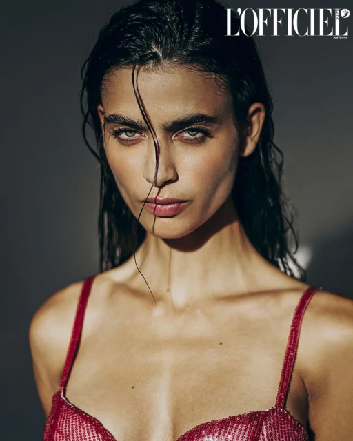 Close-up portrait of a woman with slicked-back dark hair, wearing a red sequined top, looking directly at the camera in a dimly lit studio.