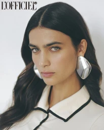 Close-up portrait of a woman with dark hair wearing large silver statement earrings and a cream-colored collared top.