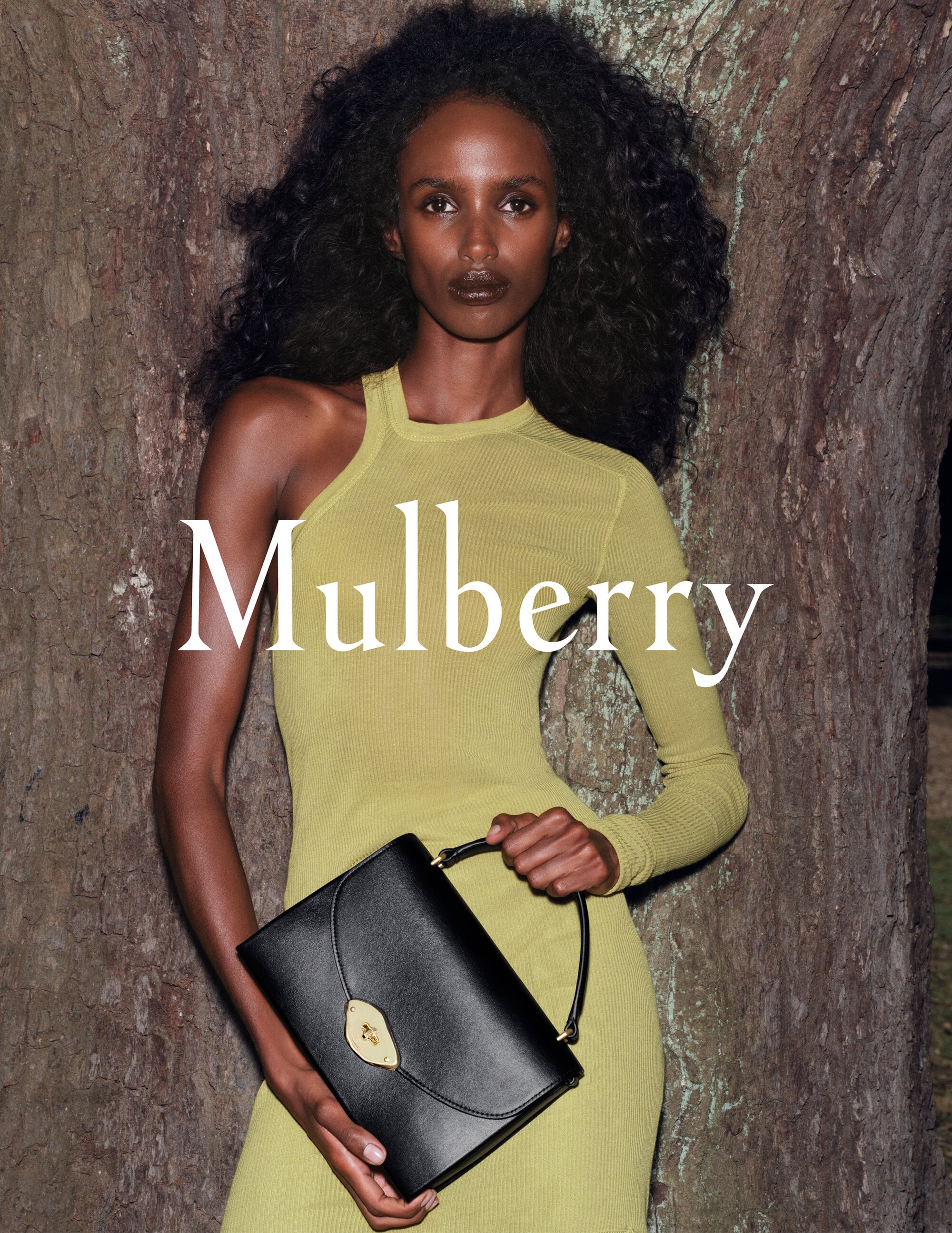 Dark-skinned fashion model poses in a fitted chartreuse dress holding a black leather Mulberry handbag, against a tree trunk background.