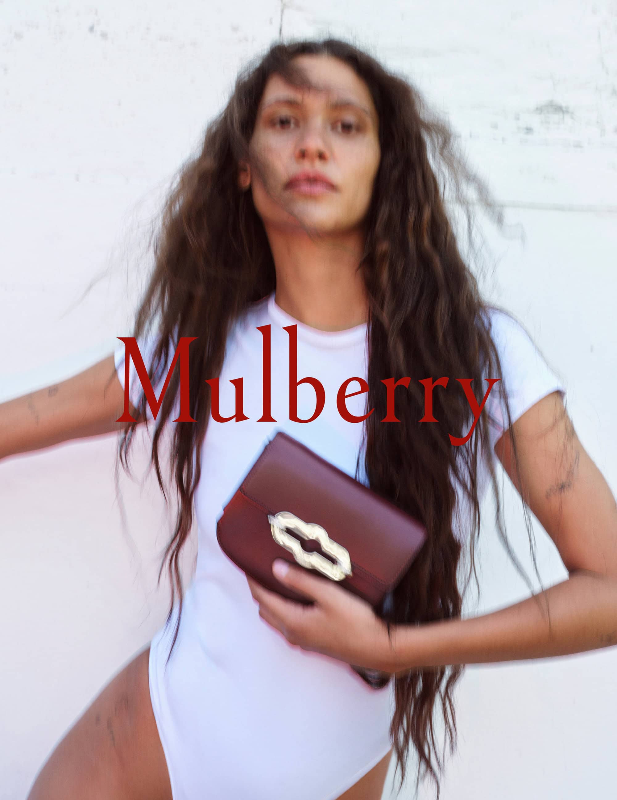 A woman with long brown hair poses indoors holding a dark red leather handbag with a metallic clasp against a white wall.