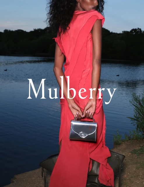Fashion shot of a model in a long, flowing red dress holding a metallic silver handbag, posing against a lake and natural backdrop.