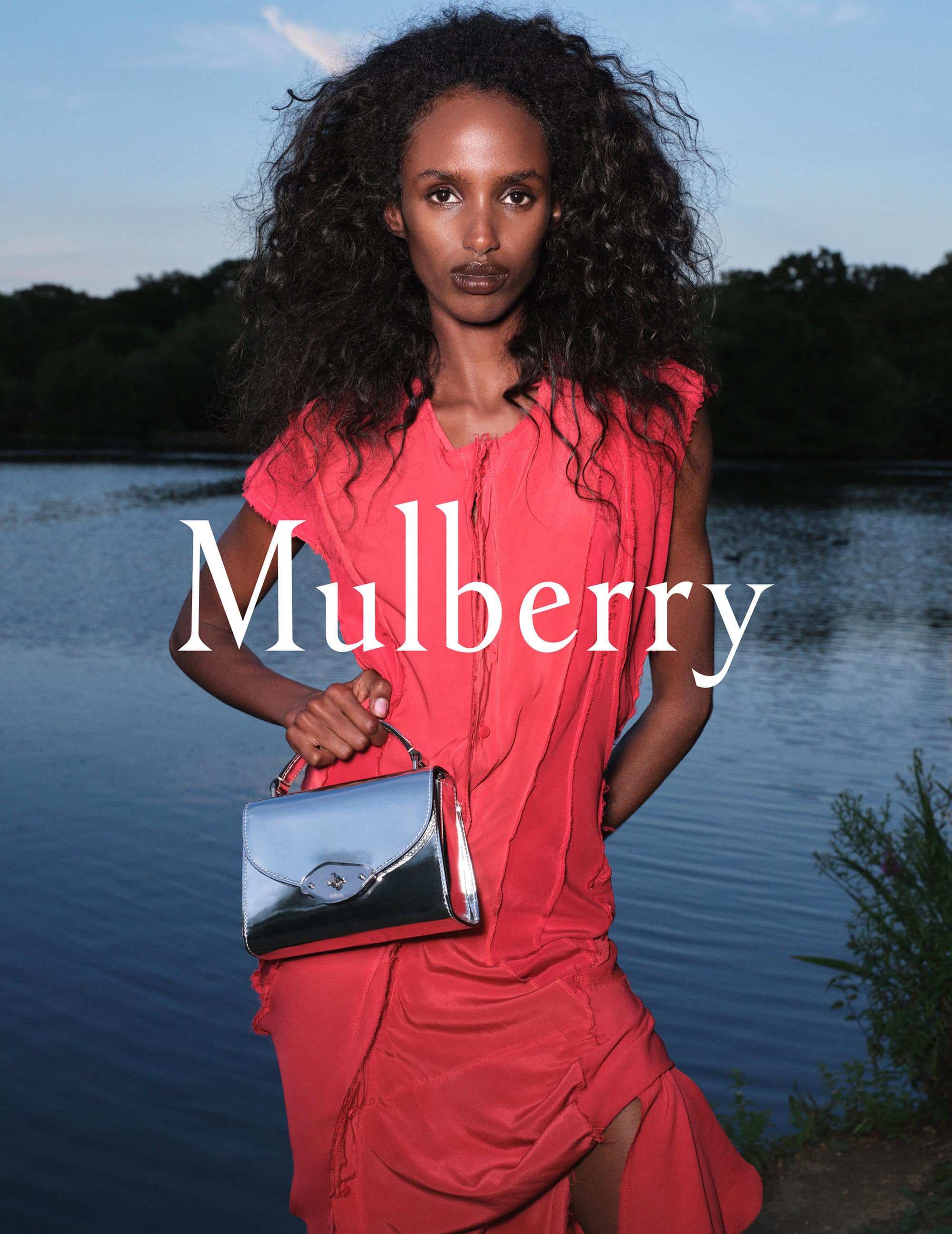 A woman poses outdoors with a silver Mulberry handbag. She is wearing a coral dress and has long, dark, curly hair with trees and water in the background.