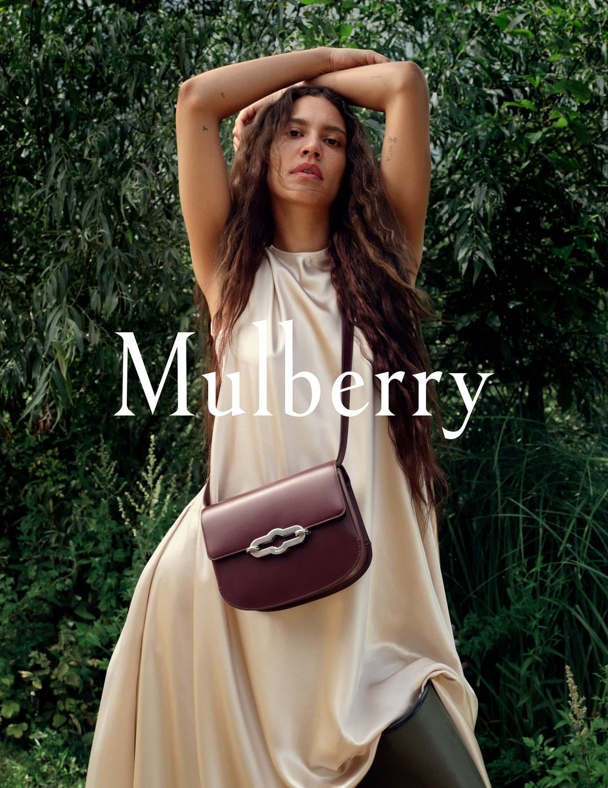 Woman in flowing dress poses with Mulberry bag in a lush, green outdoor setting, showcasing fashion and nature.