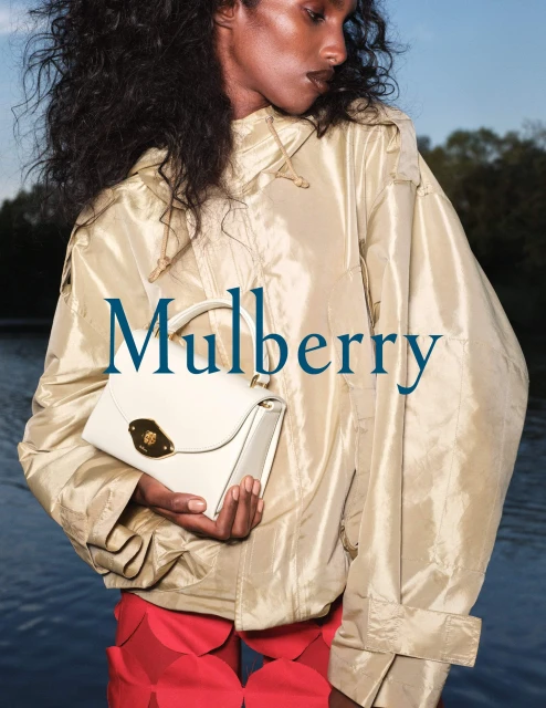 Woman presents a cream-colored handbag in a fashion setting, wearing a beige jacket and red shorts. Water is visible in the background.