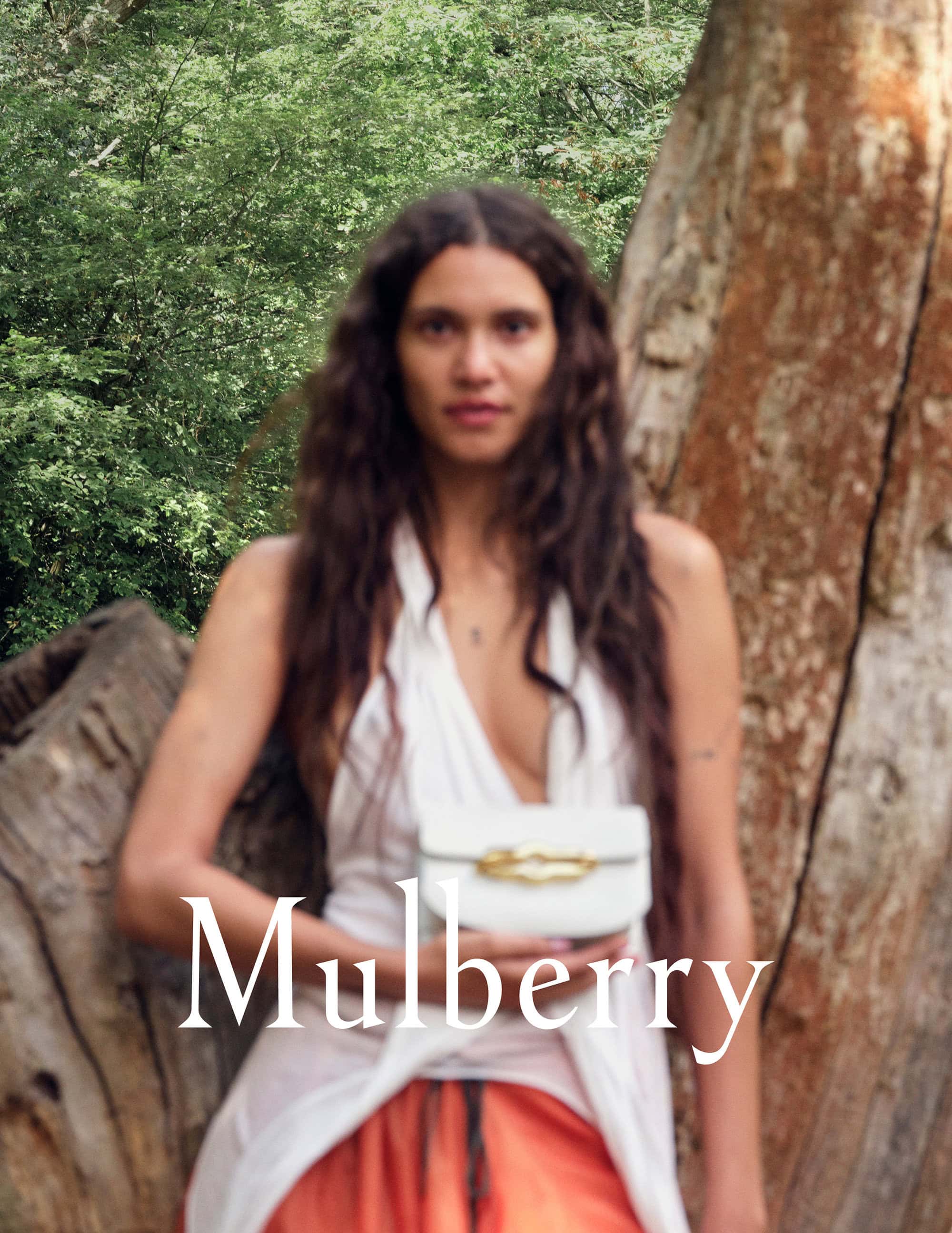 Woman in flowing white dress and orange skirt holds a white Mulberry handbag outdoors, posed against a tree with lush green foliage.
