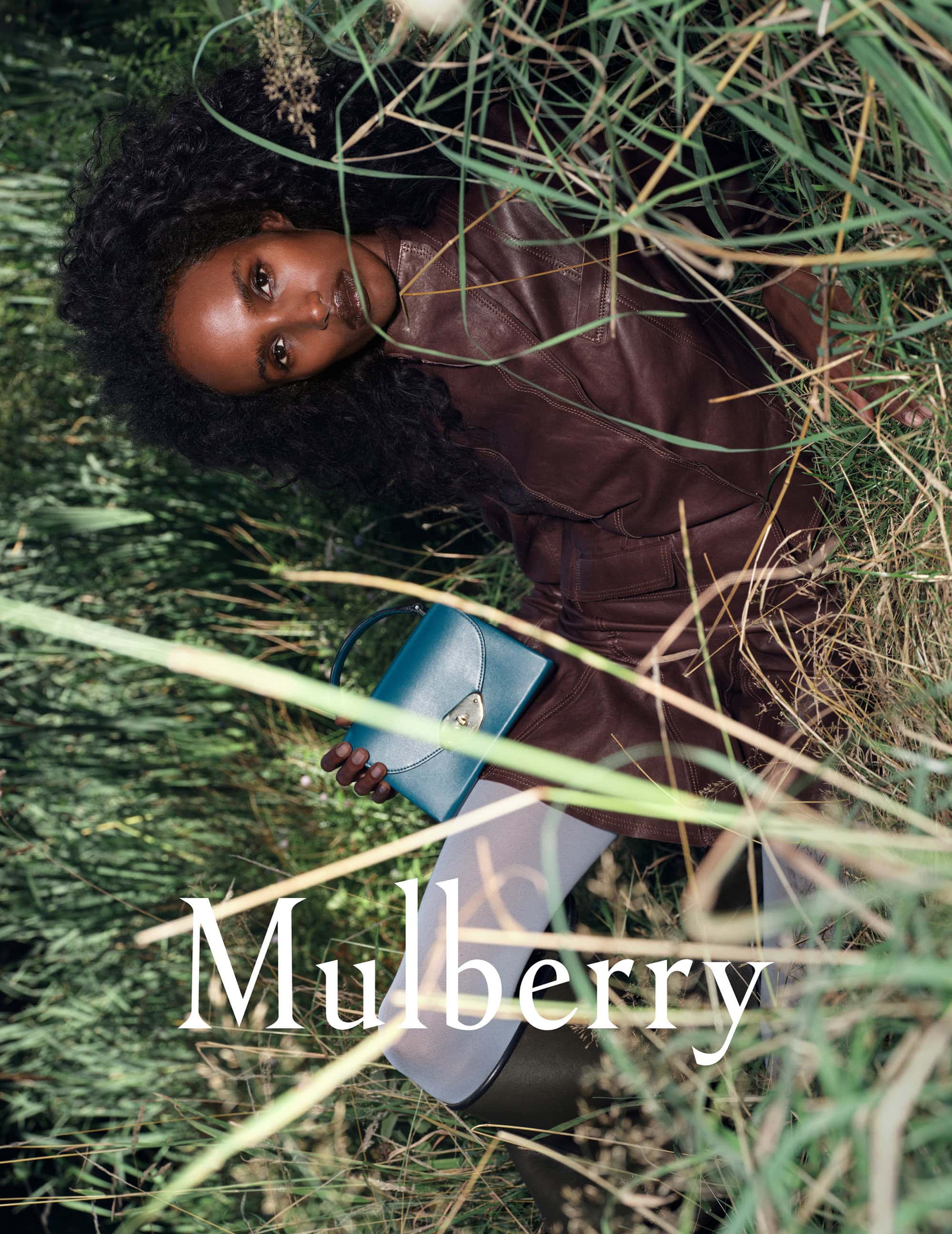 Dark-skinned woman with curly hair in a burgundy outfit, lying in tall grass, holding a teal Mulberry handbag in a fashion setting.