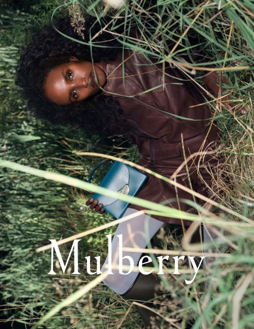 Dark-skinned woman with curly hair in a burgundy outfit, lying in tall grass, holding a teal Mulberry handbag in a fashion setting.