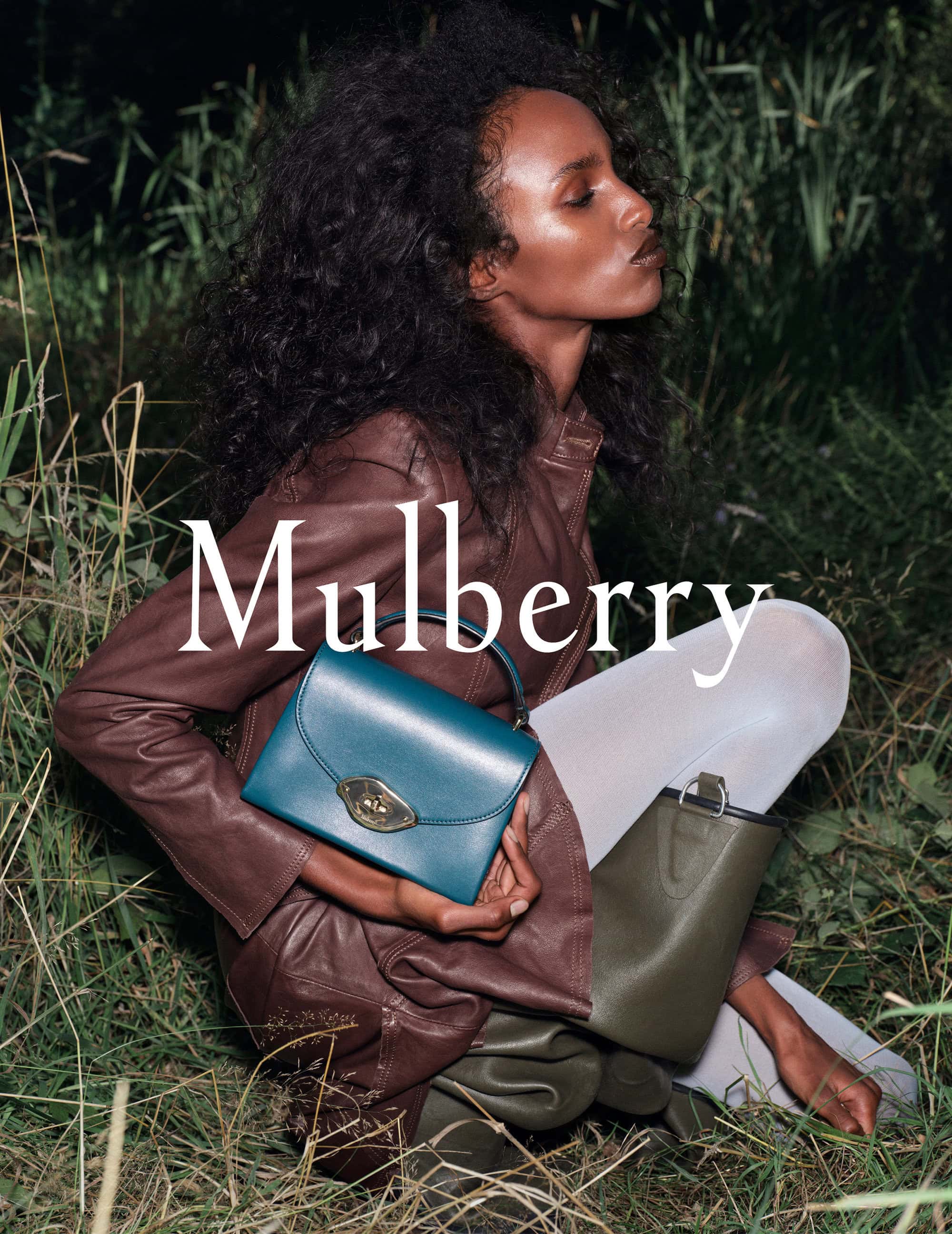 Fashion shot of a woman with dark curly hair wearing a brown leather jacket and holding a teal handbag in a grassy field.