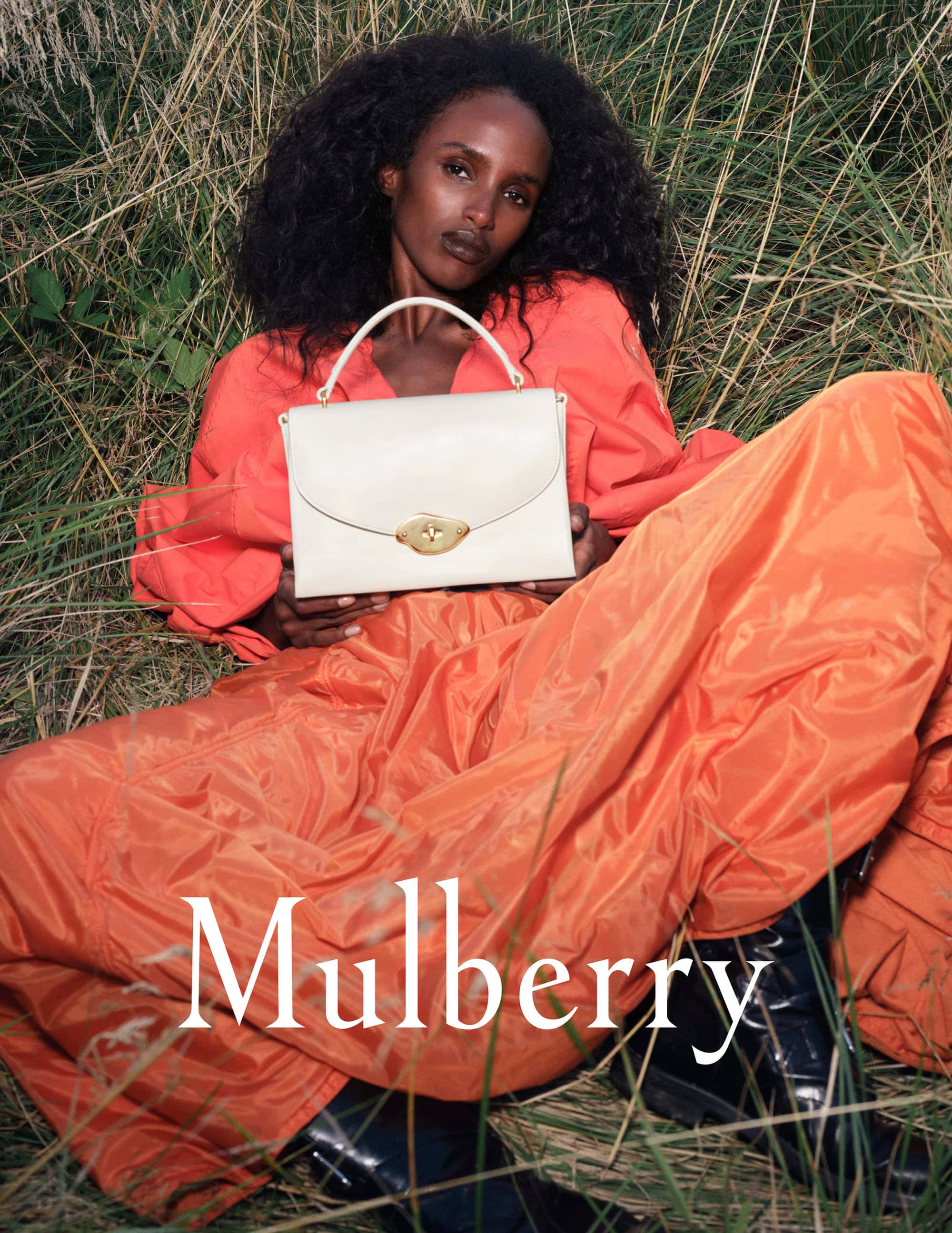 Dark-skinned female model poses with a cream leather handbag in tall grass, wearing orange blouse and skirt. Bag by Mulberry.