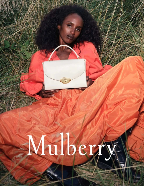 Dark-skinned female model poses with a cream leather handbag in tall grass, wearing orange blouse and skirt. Bag by Mulberry.