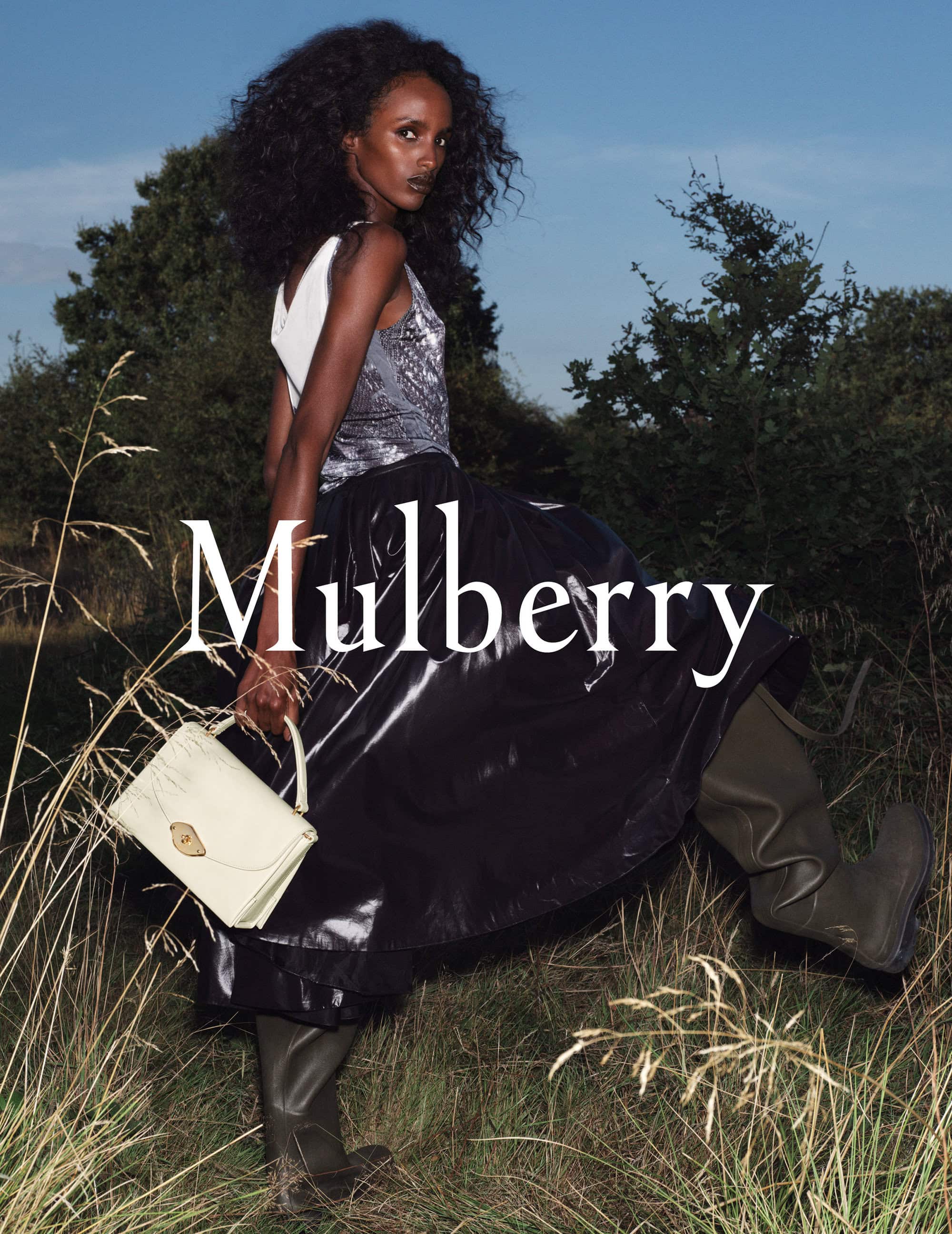 Elegant outdoor fashion portrait of a dark-skinned woman wearing a stylish top, dark skirt, and boots, holding a light-colored handbag.