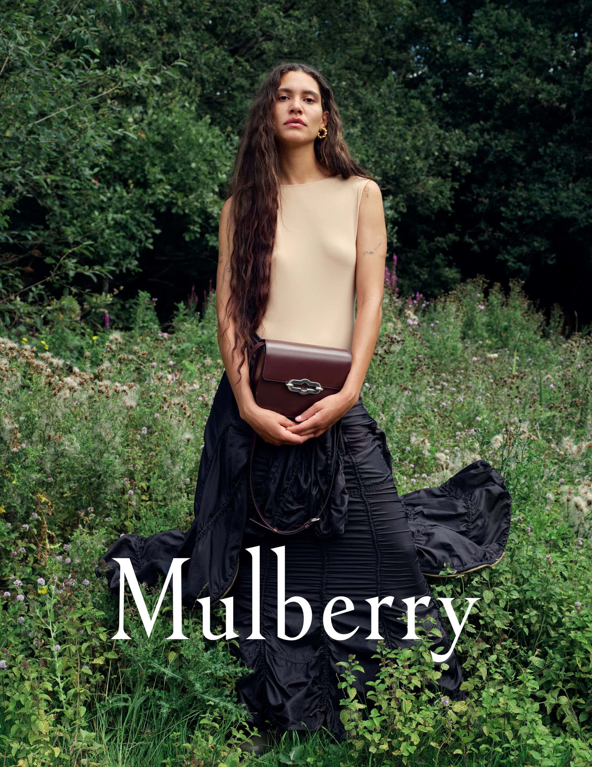 A woman with long brown hair stands in an outdoor field, wearing a light-colored top and black skirt, holding a brown leather purse.