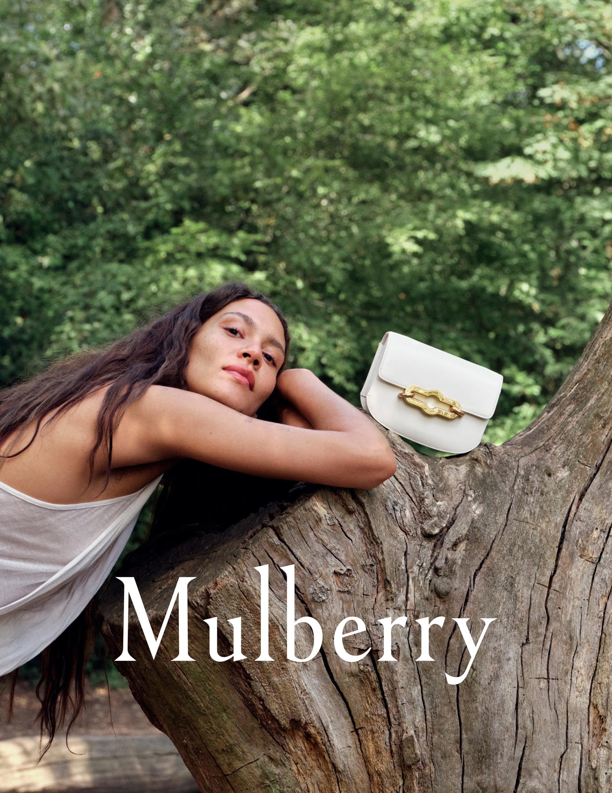 Woman rests on a fallen tree log with a white leather purse featuring gold hardware in front of a blurred green forest backdrop.