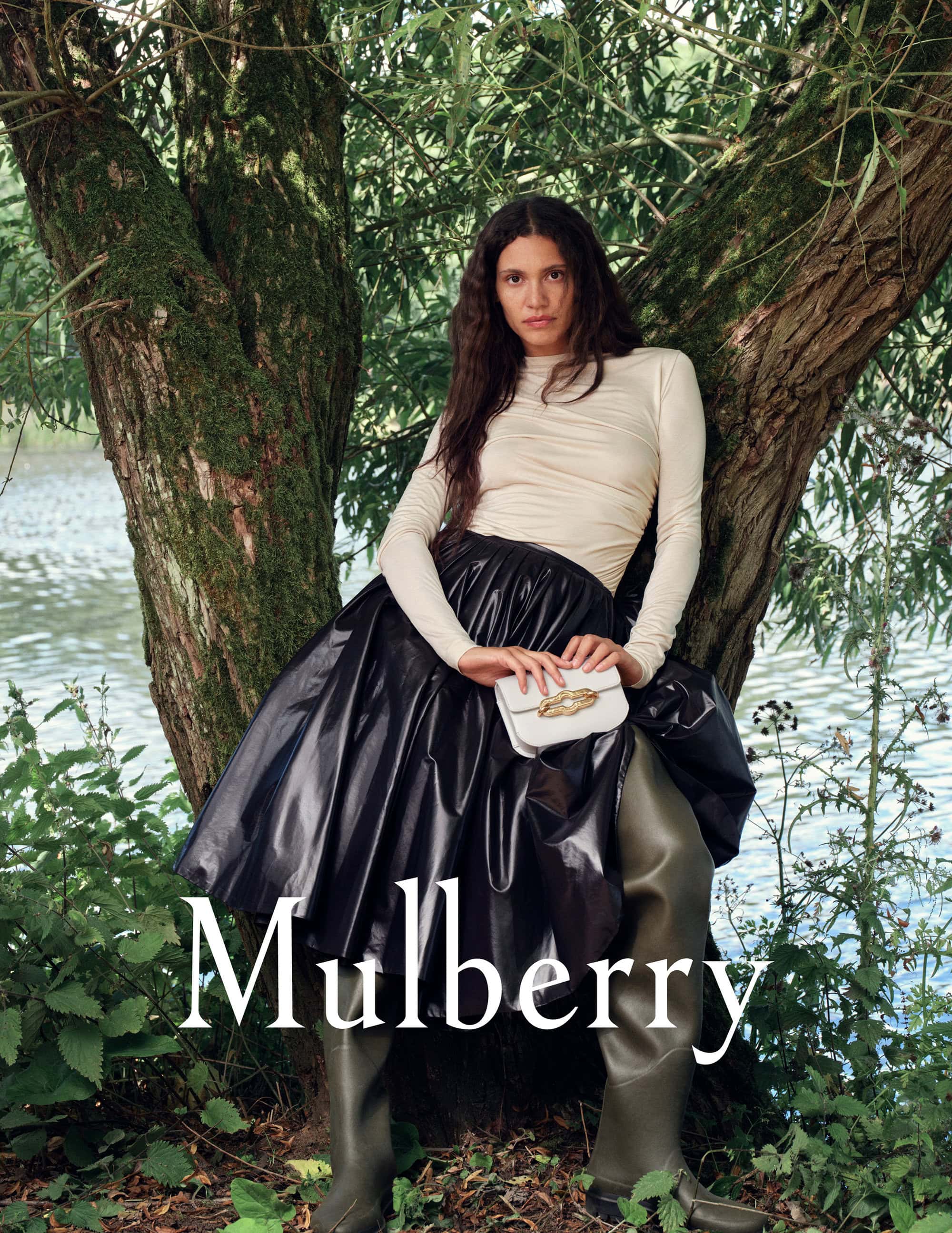 Fashion shot featuring a woman in a draped top, shiny skirt, and boots, posing against a tree with a river in the background.