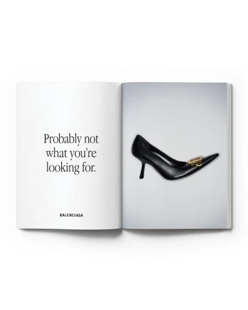 Black leather high-heel pump with gold logo on a magazine page, offset by contrasting text against a light gray backdrop.
