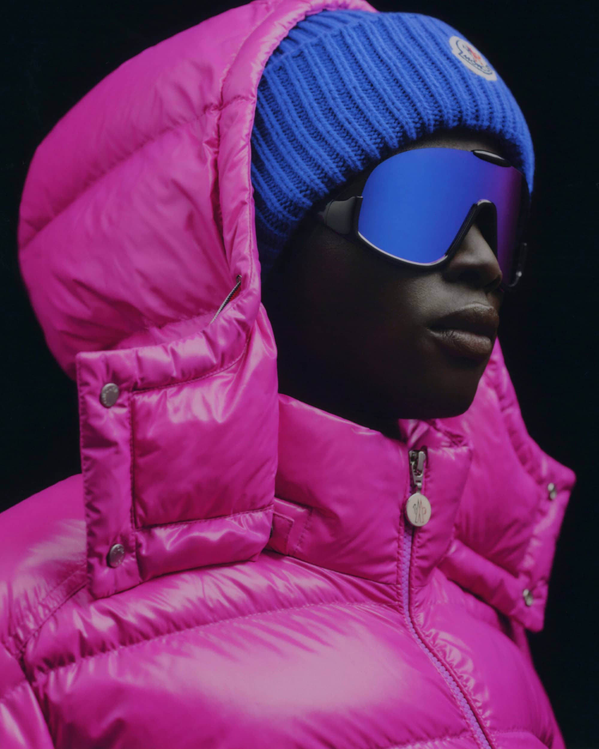 Dark-skinned person wearing a hot pink puffer jacket, blue beanie, and mirrored sunglasses against a dark background. Fashion advertisement.