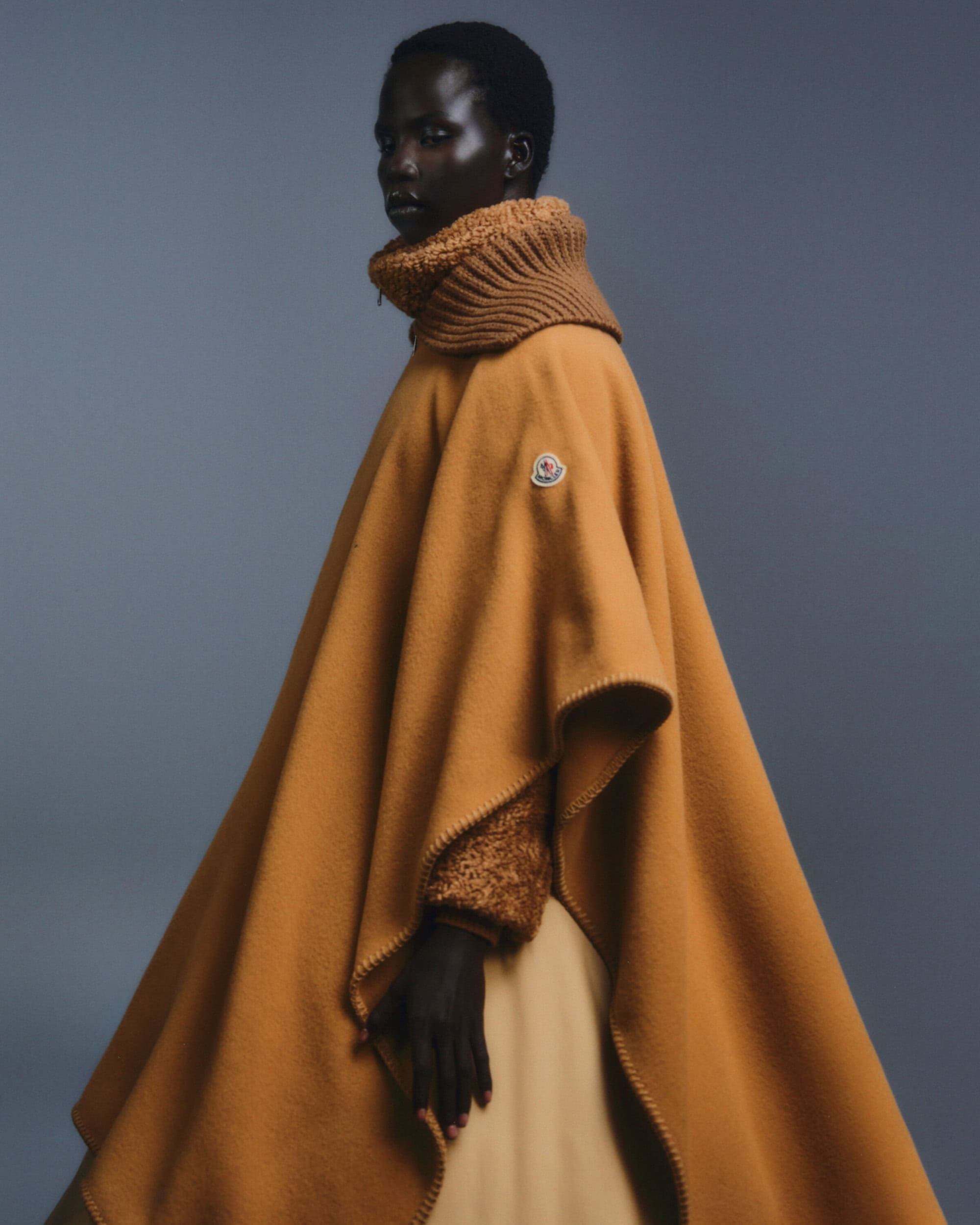 Fashion editorial shot of a Black model wearing an orange wool poncho with a ribbed turtleneck against a grey gradient background.