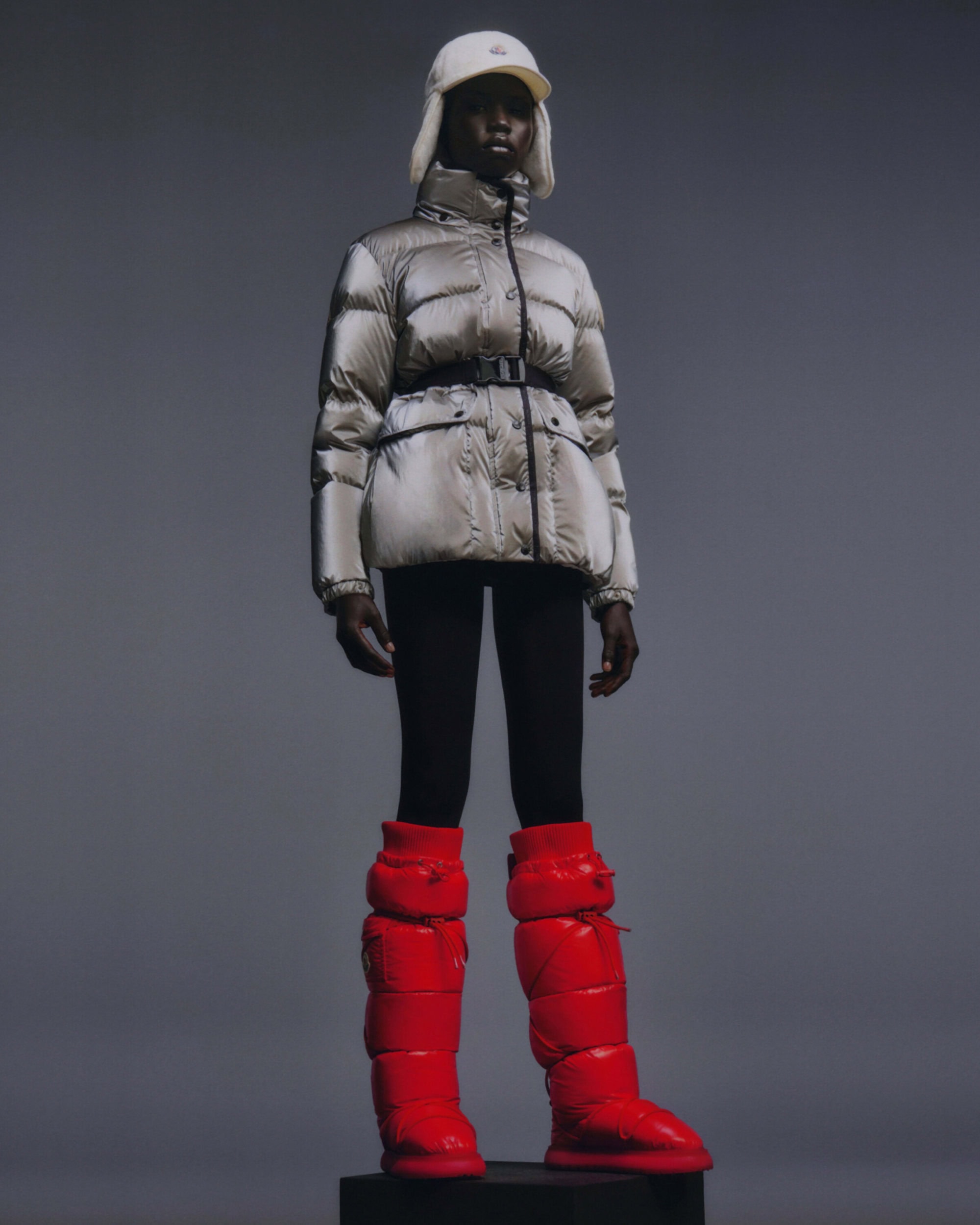 Fashion shot: a Black model posing in a grey puffer jacket, black leggings, and red puffer boots against a grey backdrop. 
