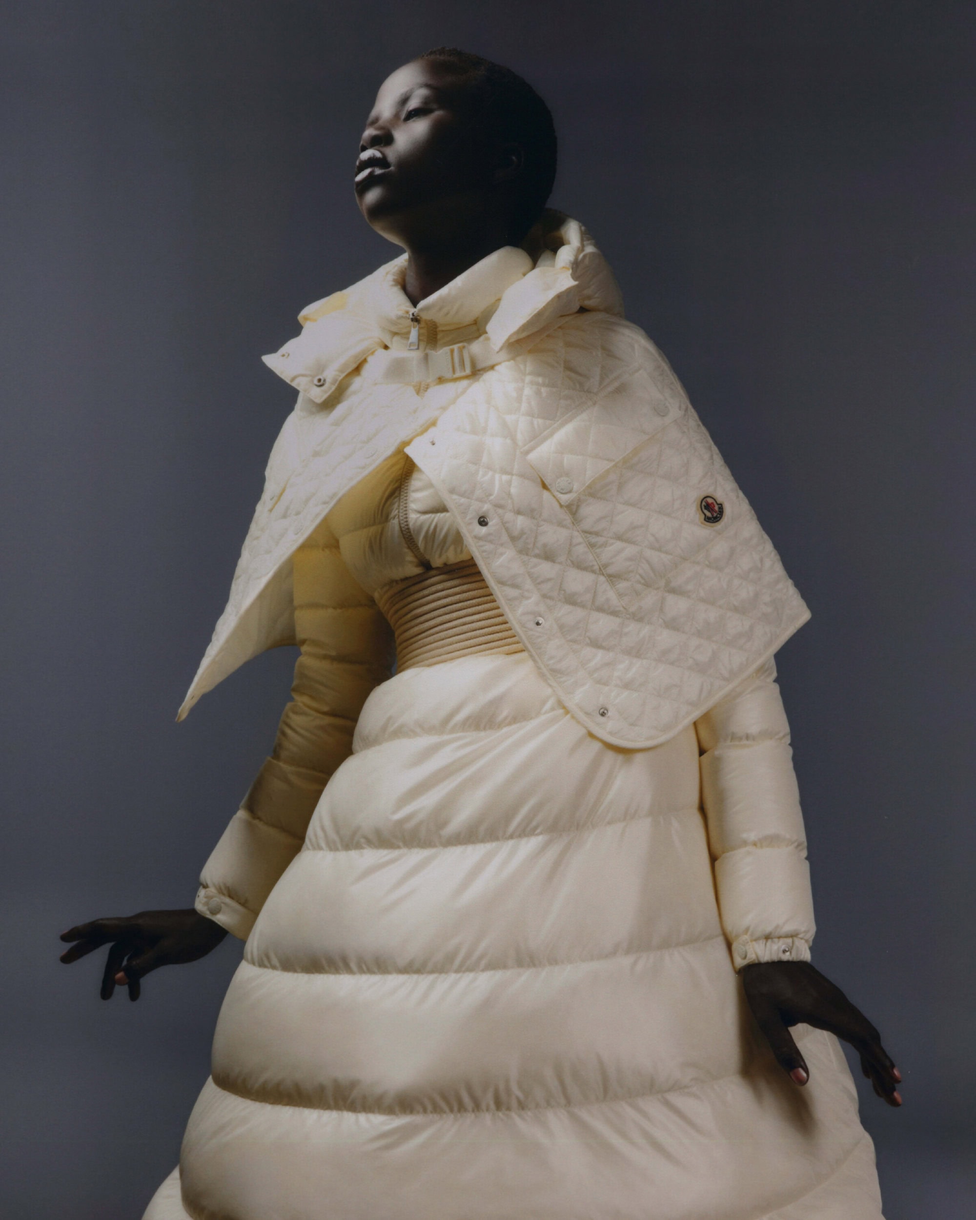 A dark-skinned woman poses in a studio wearing a white quilted coat dress and cape, looking upwards against a grey backdrop in a fashion shot.