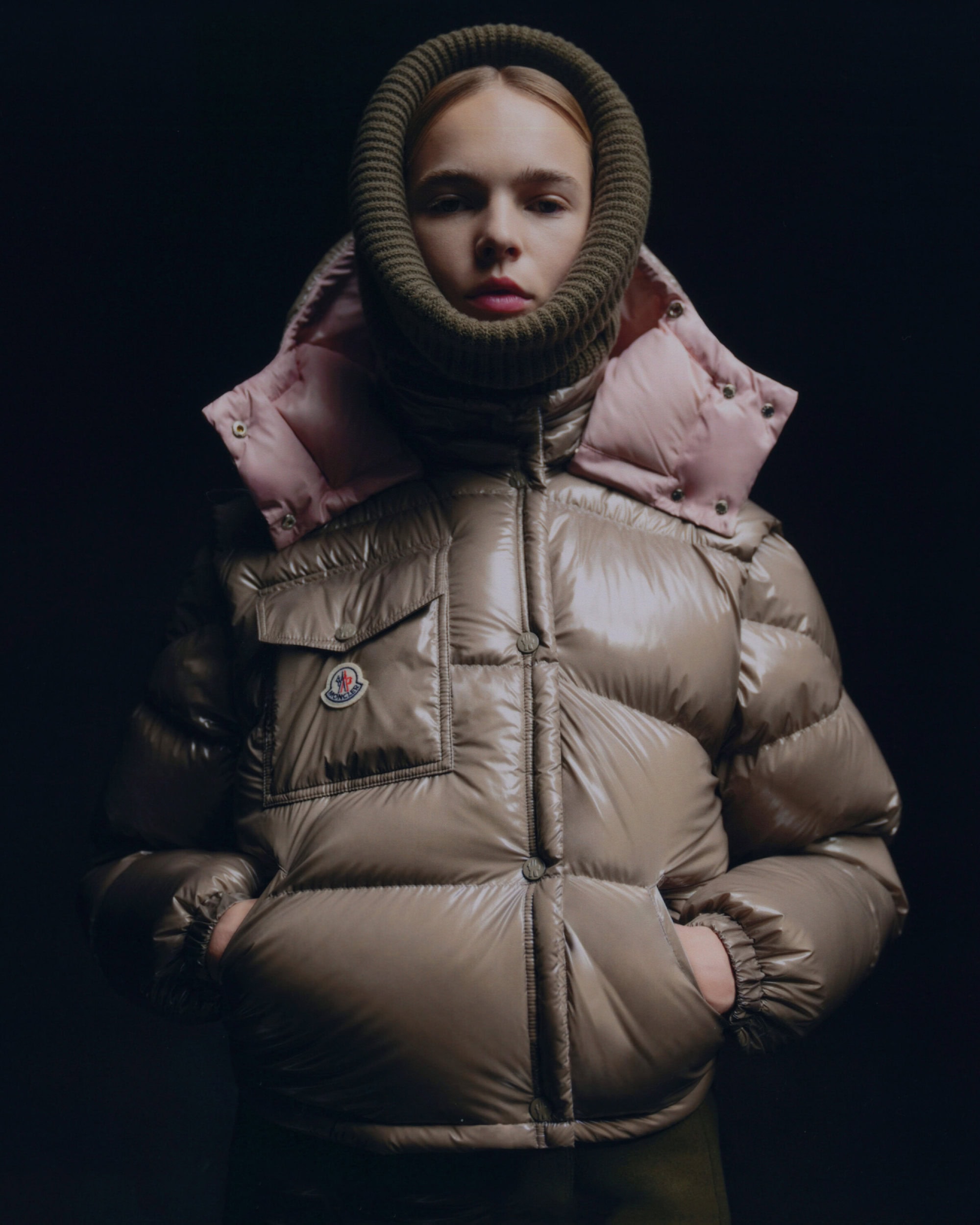 Close-up studio shot of a fair-skinned woman wearing a khaki-toned Moncler down puffer jacket with a knitted balaclava and a pink hood.