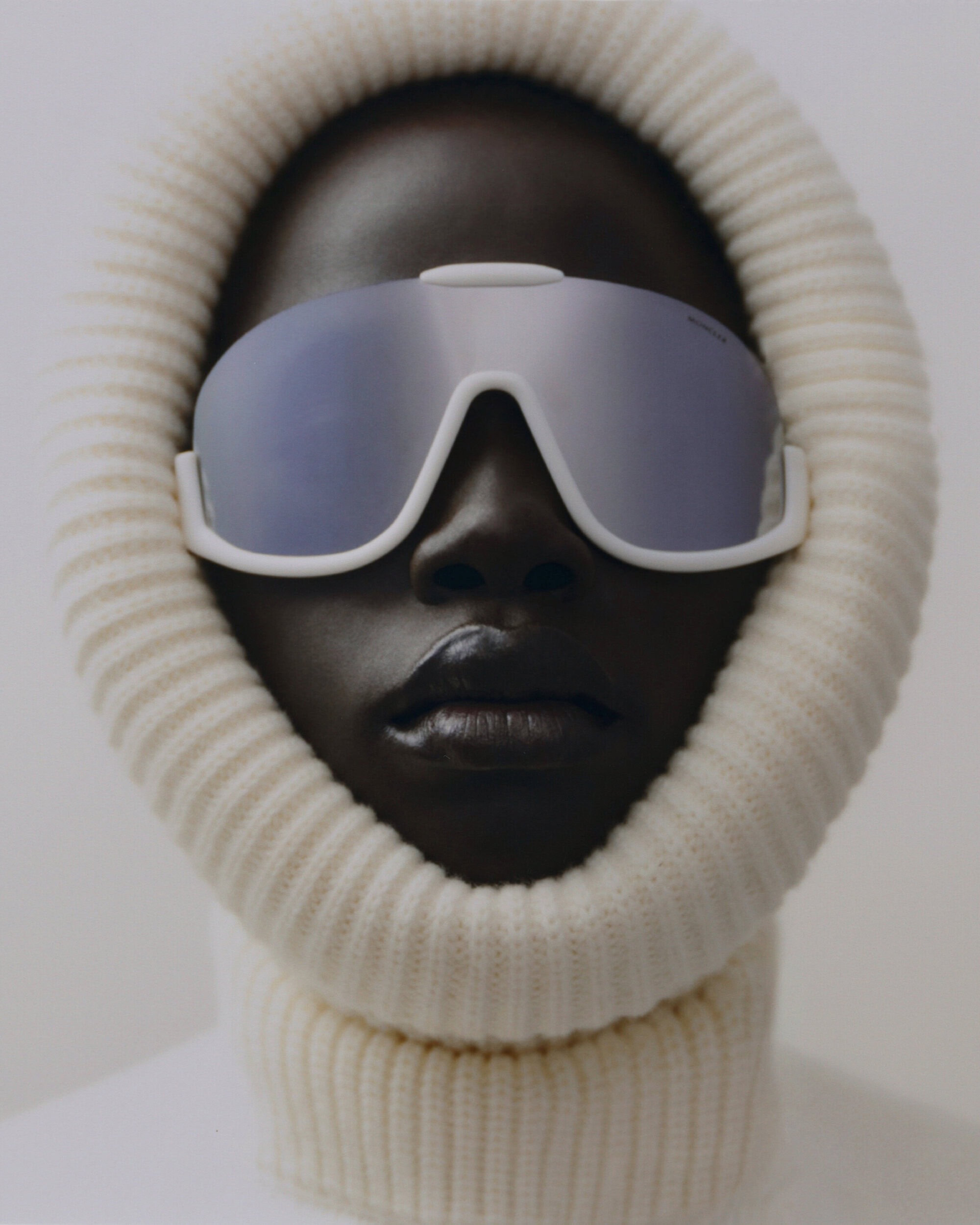 Close-up studio shot of a Black woman wearing white ski goggles and a knit balaclava against a white backdrop, capturing a minimalist aesthetic.