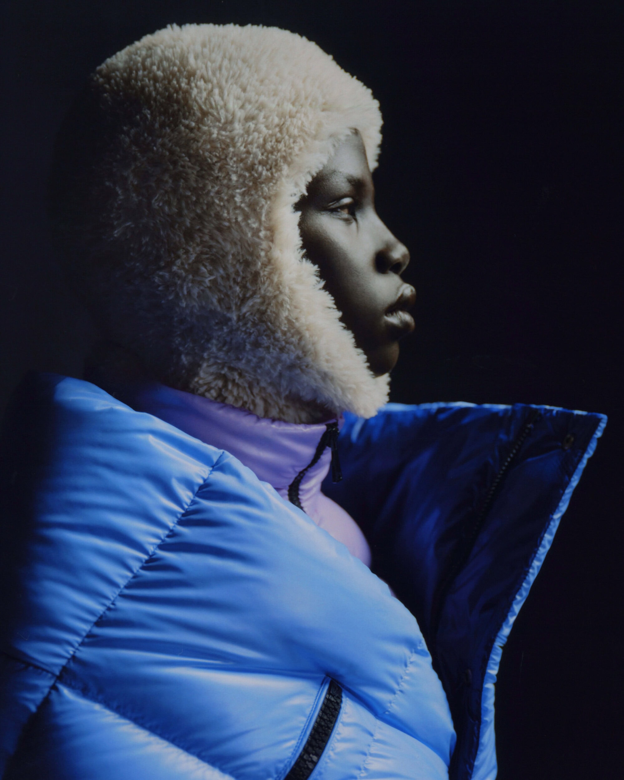 Profile shot of a person with dark skin wearing a fluffy hat and blue puffer jacket against a dark background in a studio setting.