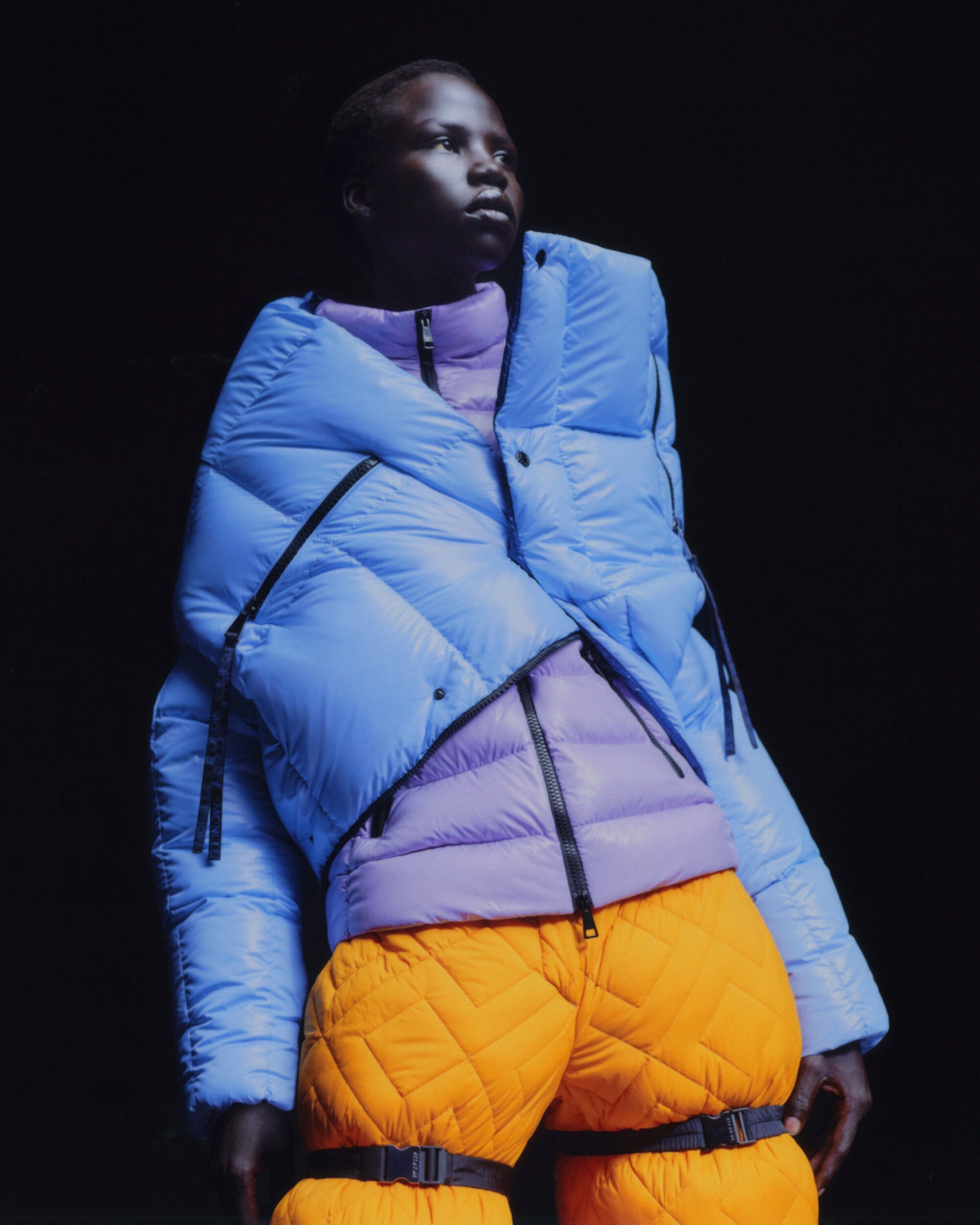 Editorial fashion shot of a Black model wearing a light blue down jacket, lilac turtleneck, and yellow quilted pants against a dark background.