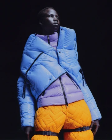 Model wearing a blue puffer jacket, purple quilted inner layer, and orange trousers against a dark background.