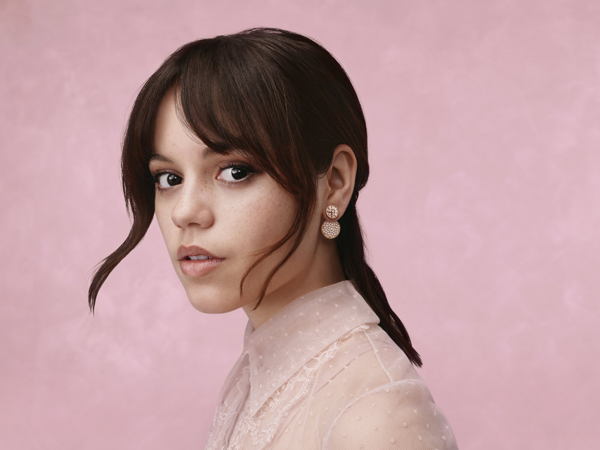 Portrait of an adult woman with long brown hair, wearing sheer blush pink blouse against soft pink background, looking at camera.