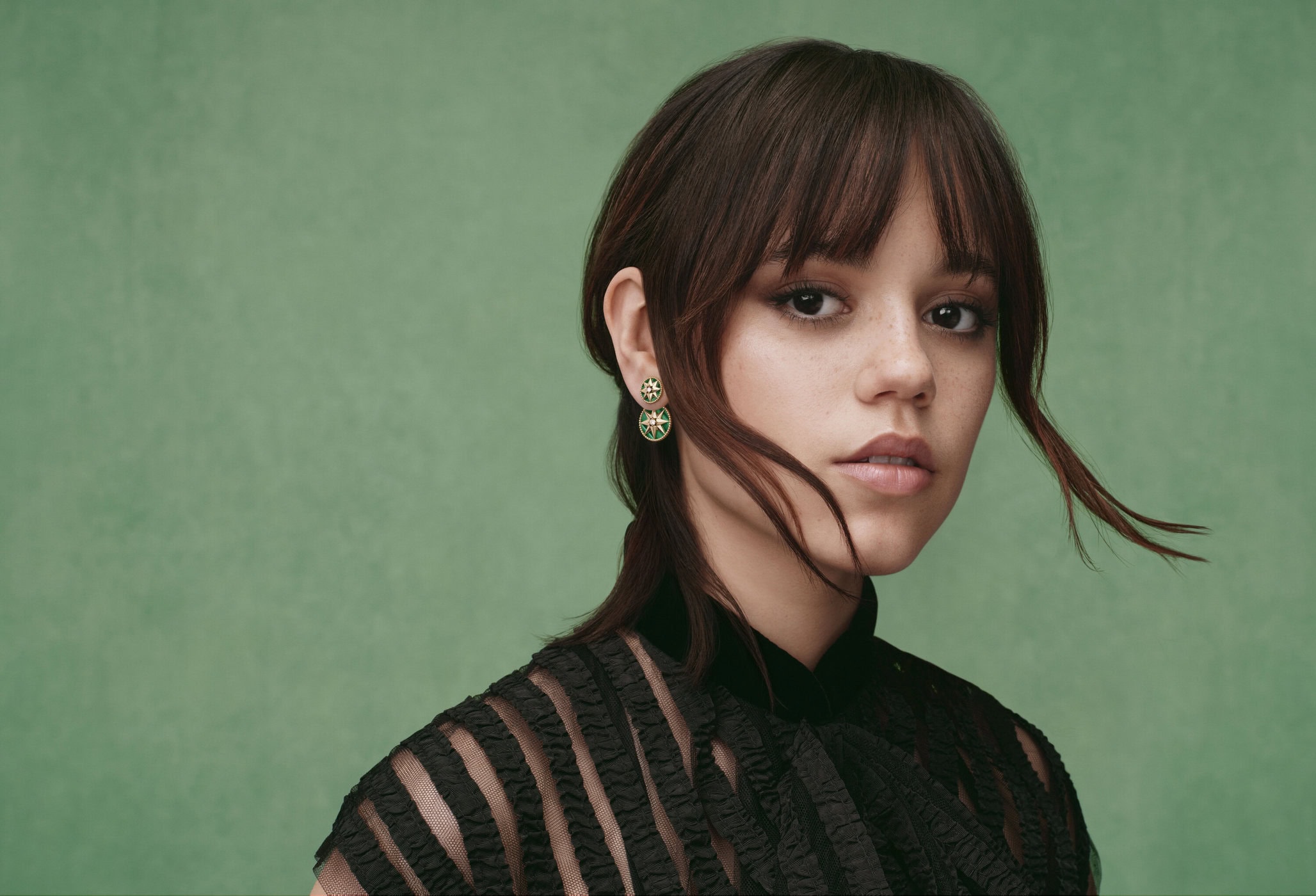 Portrait of a young adult woman with dark hair, wearing a black top and round earrings, against a solid green studio backdrop.