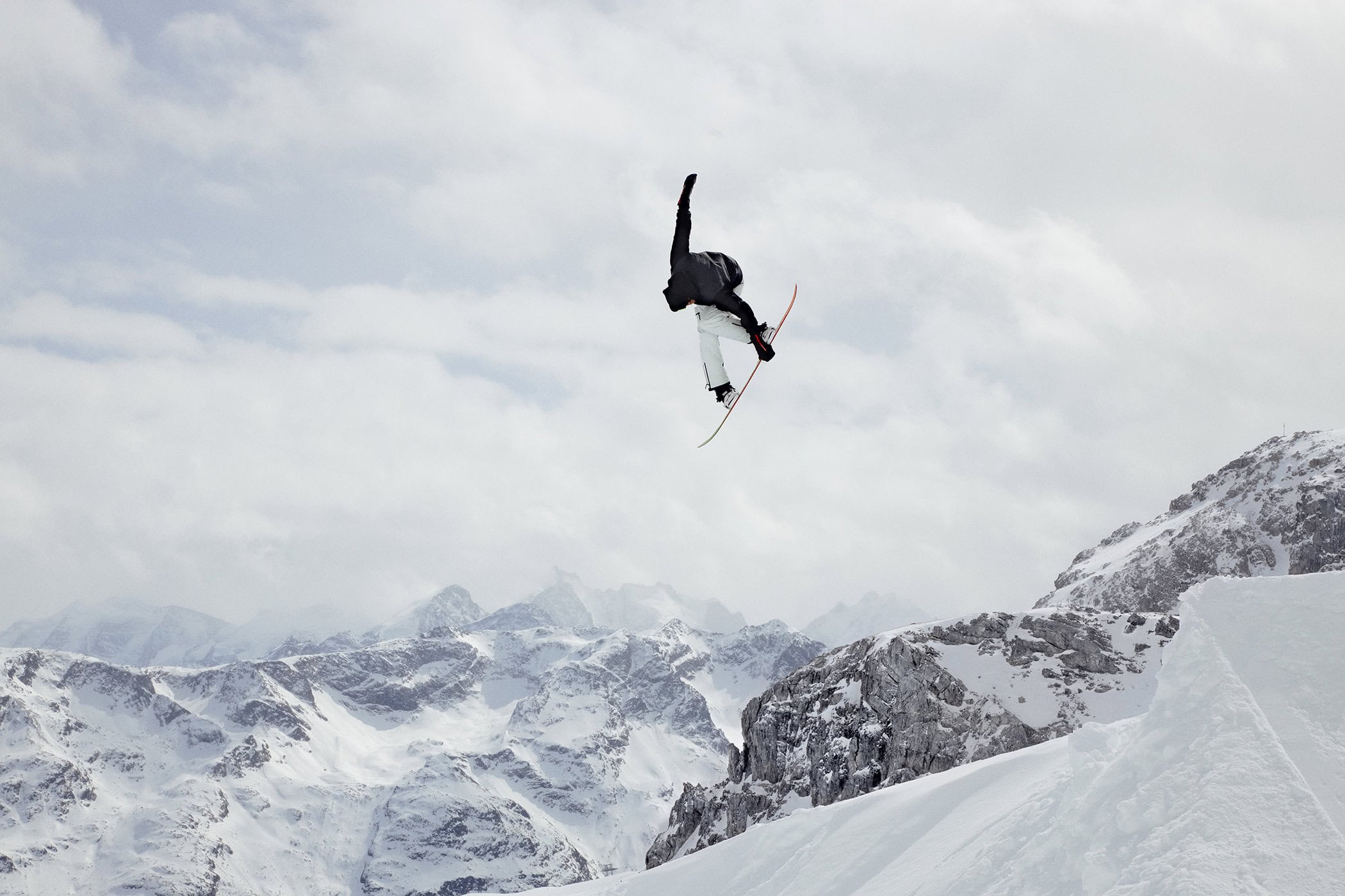 A snowboarder performs a trick against a snowy mountain backdrop under a cloudy sky, showcasing winter sports and alpine scenery.
