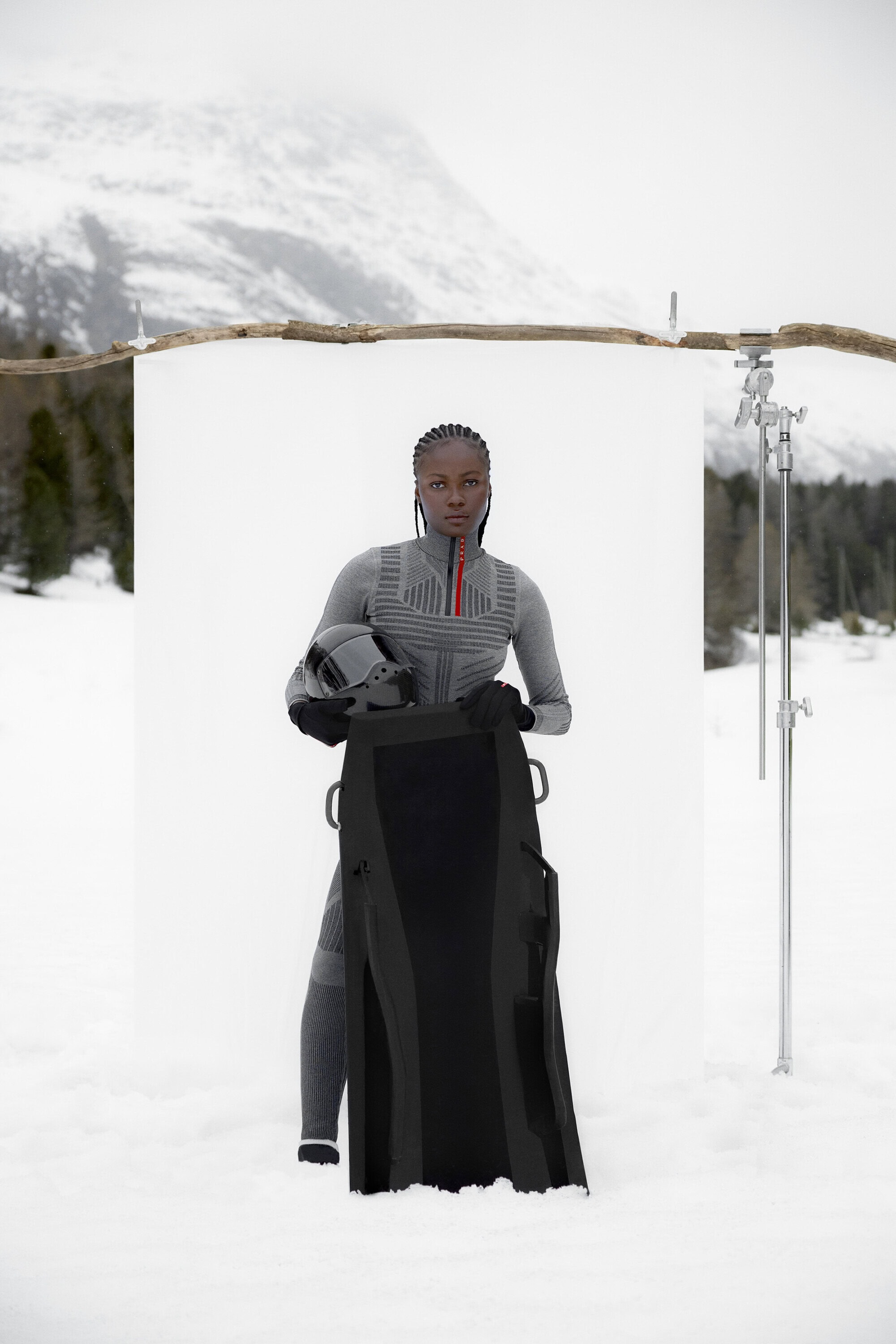 A female model stands in a snowy, mountainous landscape wearing grey athletic wear while holding a helmet and snowboard bag. 