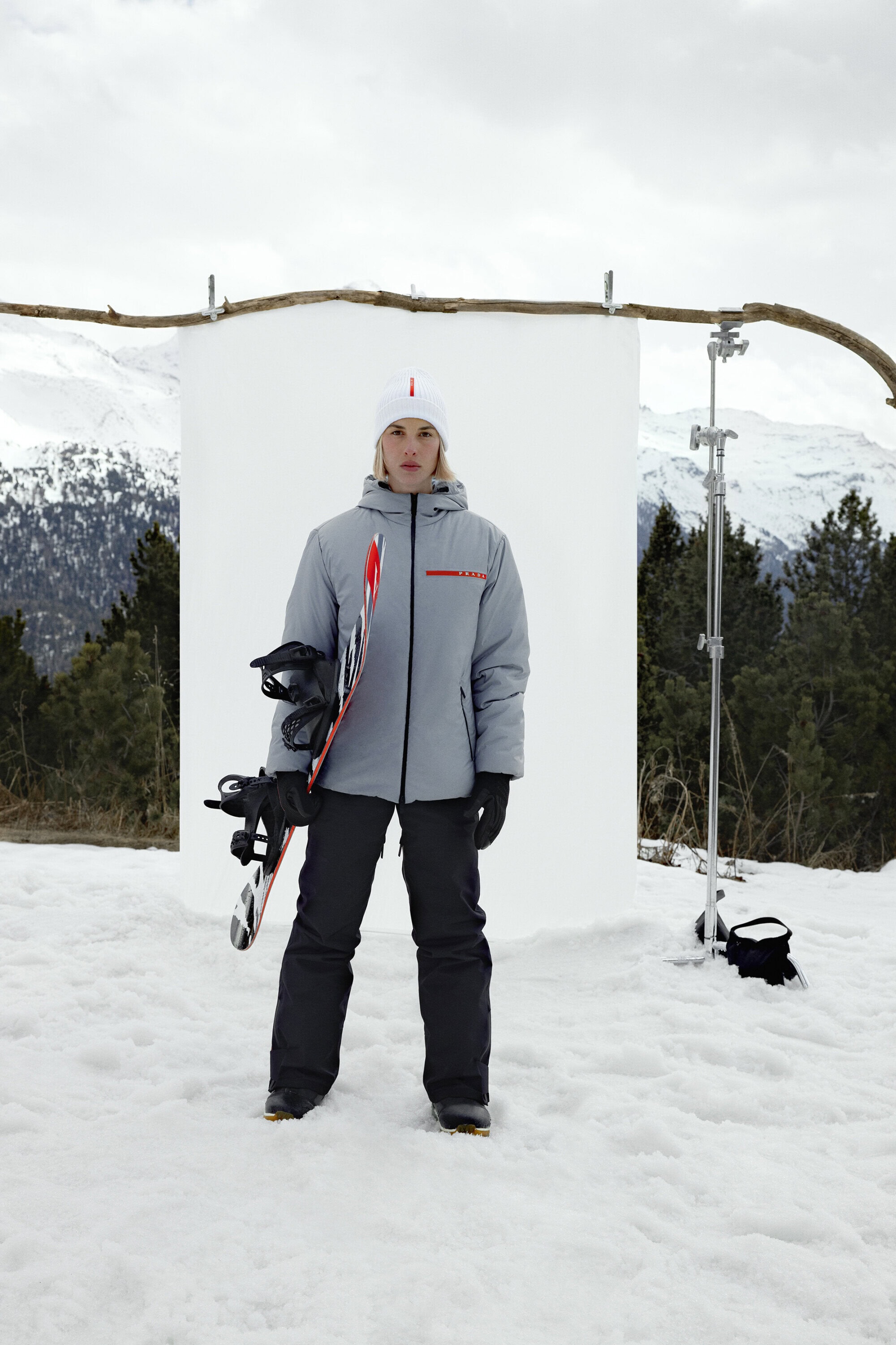 Full shot of a fair-skinned adult female standing in a snowy outdoor setting, holding a snowboard against a white backdrop.