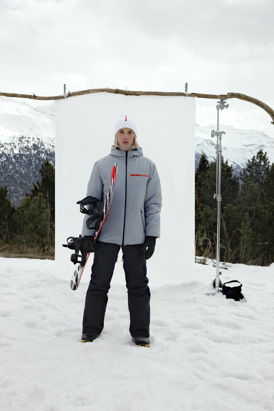 Model standing outdoors in snow wearing a grey jacket, white beanie, and dark trousers while holding a snowboard
