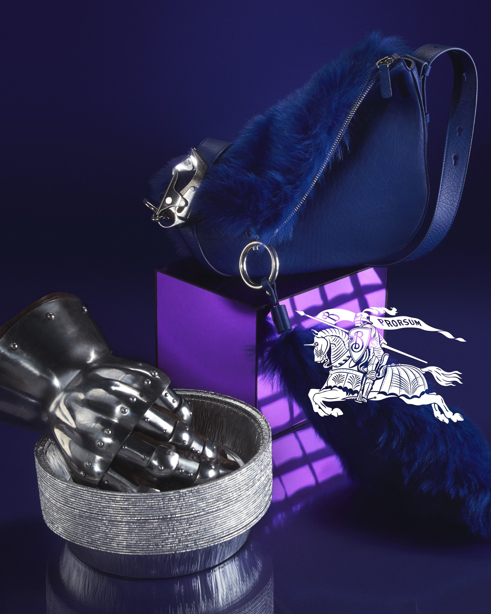 Fashion still life featuring a blue fur-trimmed handbag, silver bowl with gauntlet, and a Burberry keychain on a gradient blue background.