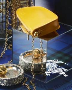 Yellow leather bag with a gold chain strap displayed atop a mirrored cube over blue tartan fabric.