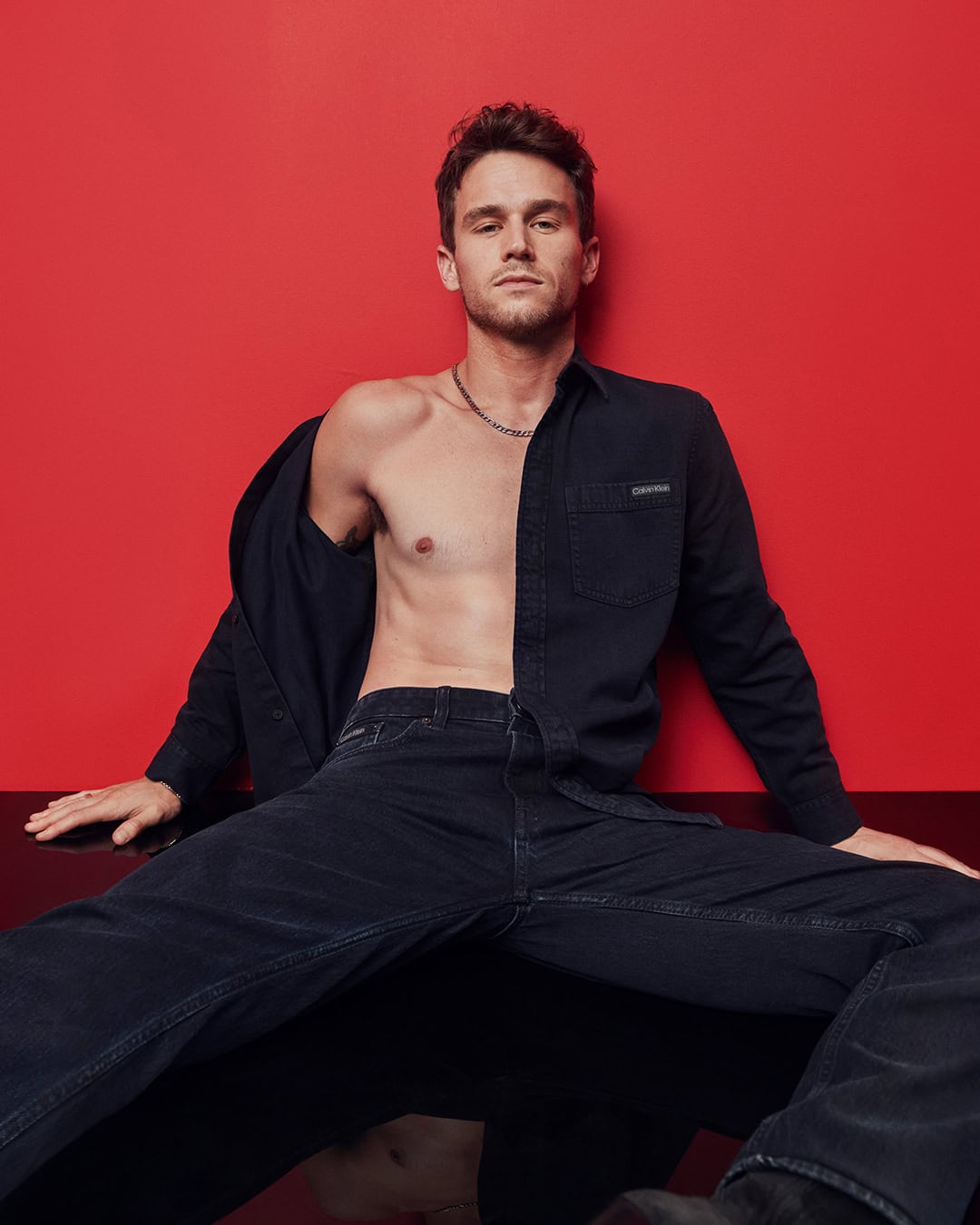 Adult male seated in a studio, wearing black denim jeans and an unbuttoned black button-down shirt against a red background.
