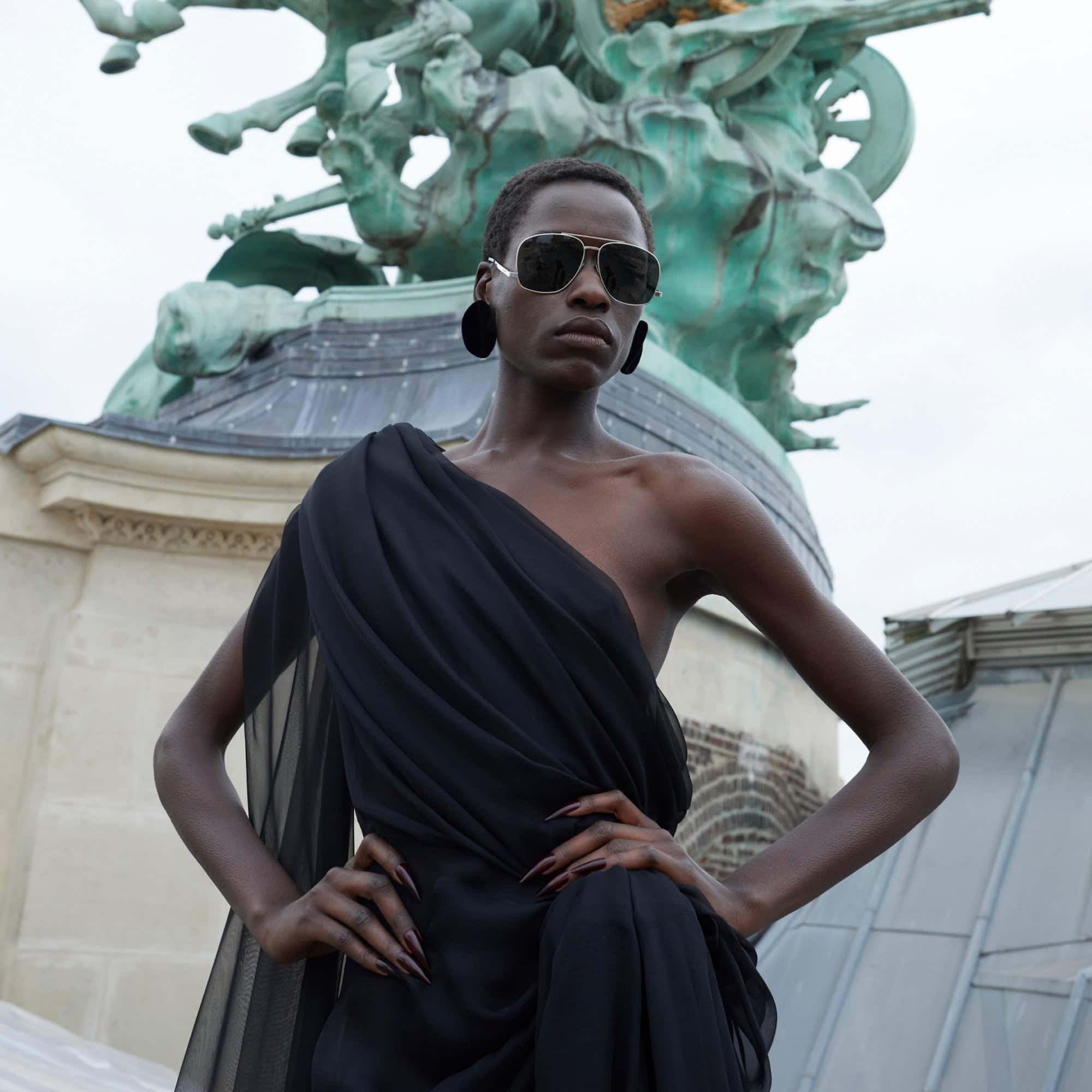 Elegant woman with sunglasses and black dress poses in front of ornate sculpture, showcasing fashion and urban sophistication outdoors.