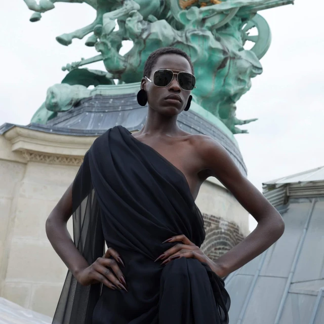 Elegant woman with sunglasses and black dress poses in front of ornate sculpture, showcasing fashion and urban sophistication outdoors.